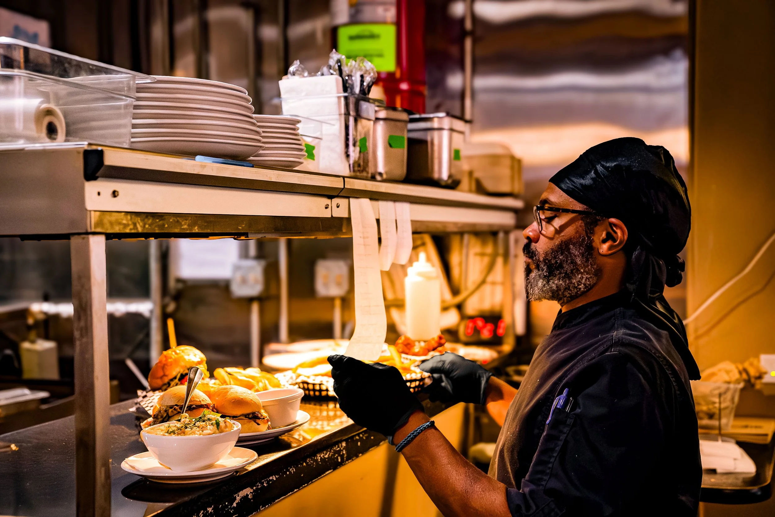 A chef with glasses, a beard, and wearing a black headscarf and gloves, preparing food at a kitchen counter filled with bowls and plates of food, with a warm light illuminating the workspace.