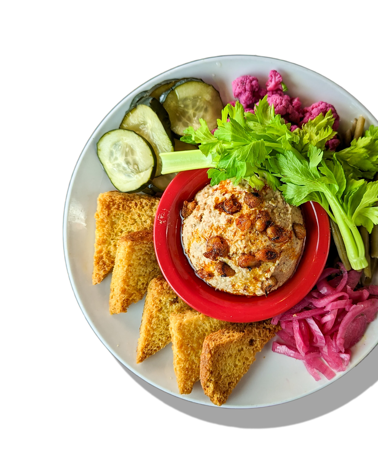 Plate of hummus with pine nuts, fresh herbs, sliced cucumbers, pickled pink onions, and toasted pita chips on a black background.