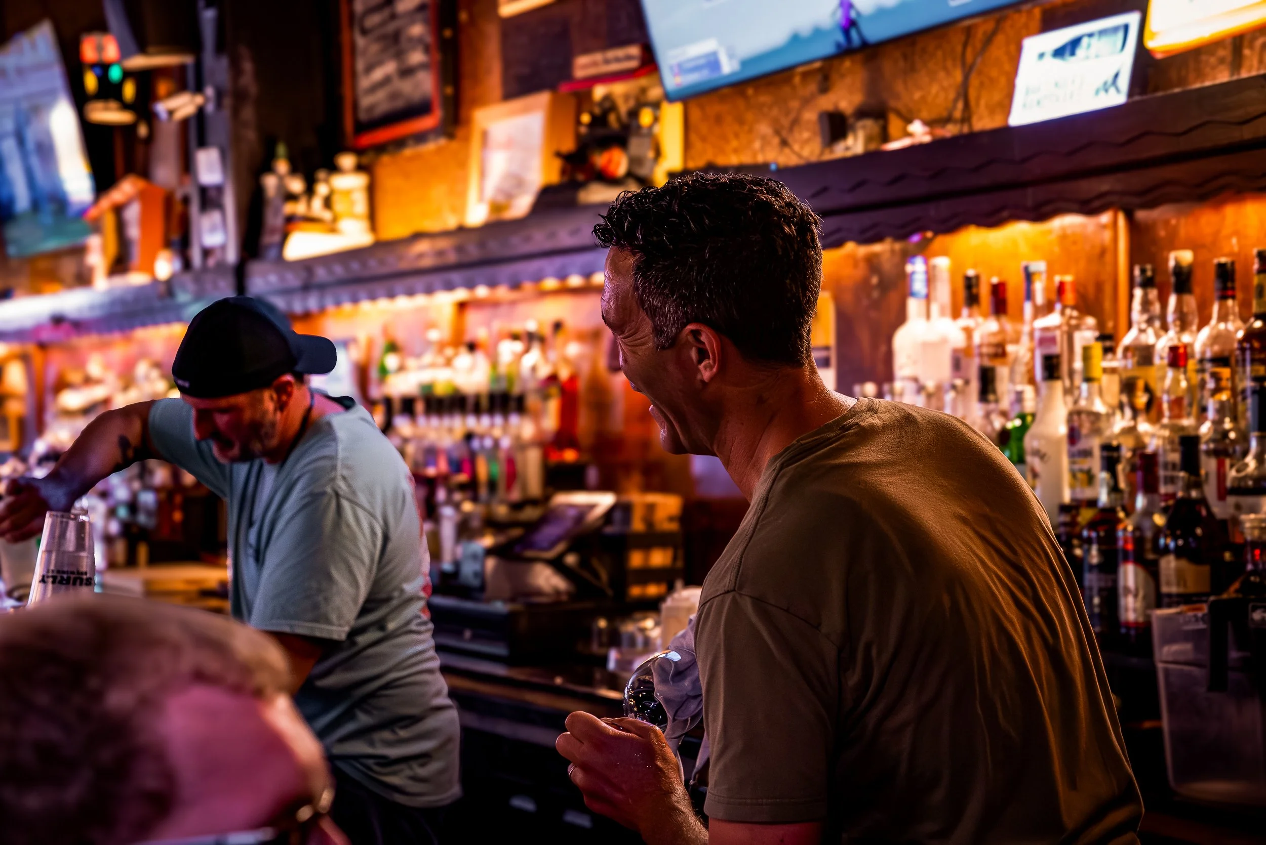 People laughing and enjoying drinks at a bar with shelves of liquor bottles and a TV screen in the background.