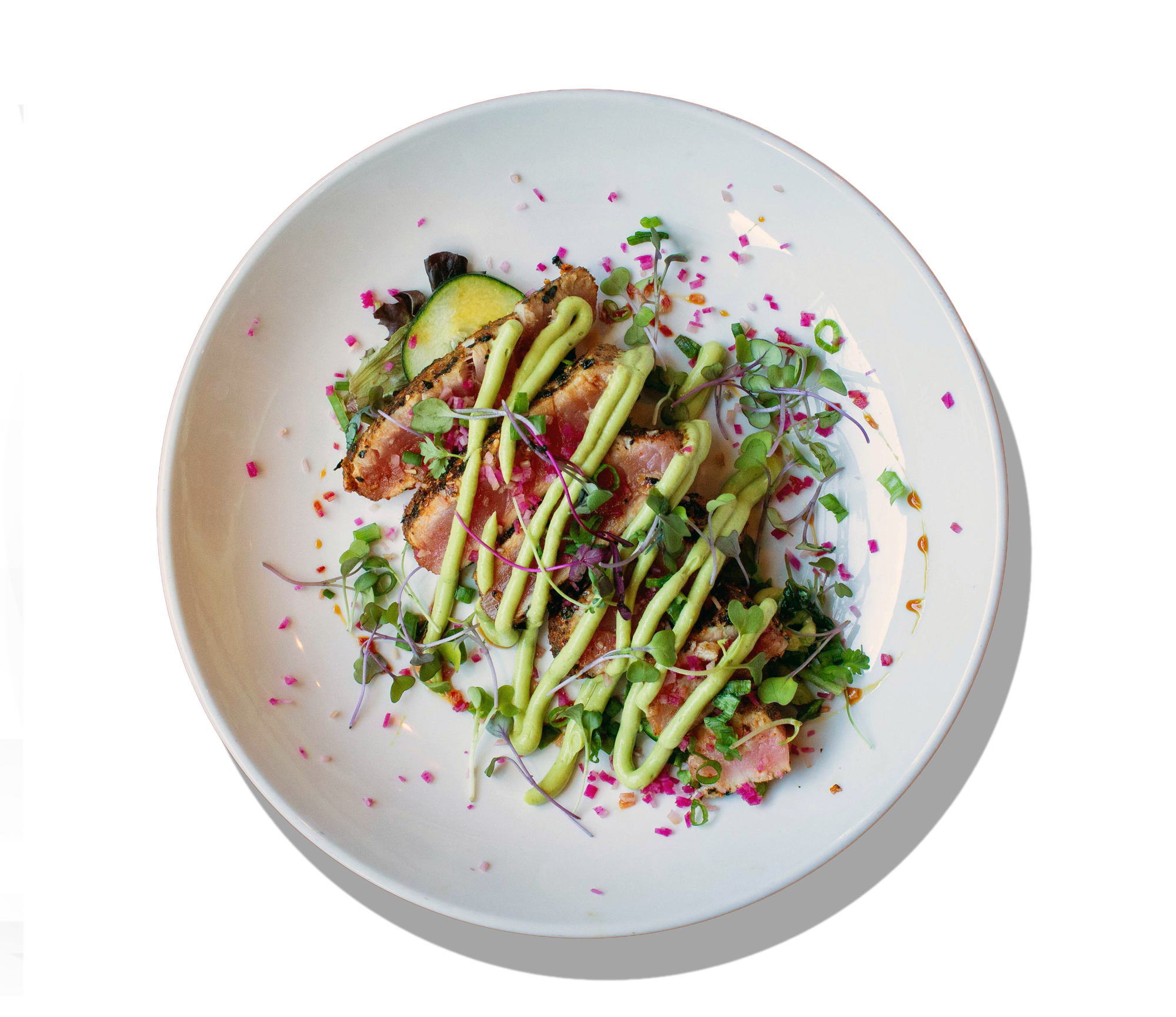 A plate of salad with sliced cucumber, microgreens, grilled meat slices, green dressing drizzle, and sprinkled pink, purple, and green garnishes on a white dish.