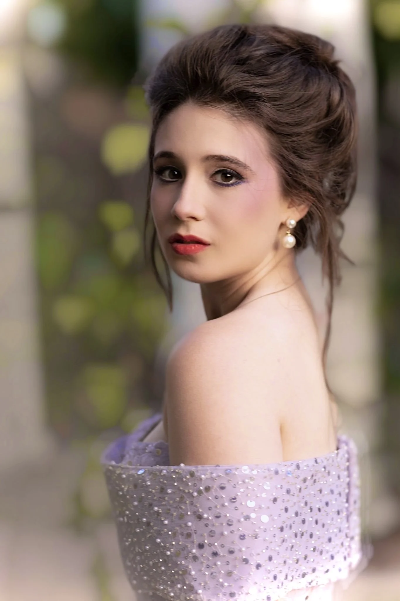 A woman with styled brown hair, wearing pearl earrings and a sparkly off-shoulder dress, looking directly at the camera with a blurred outdoor background.