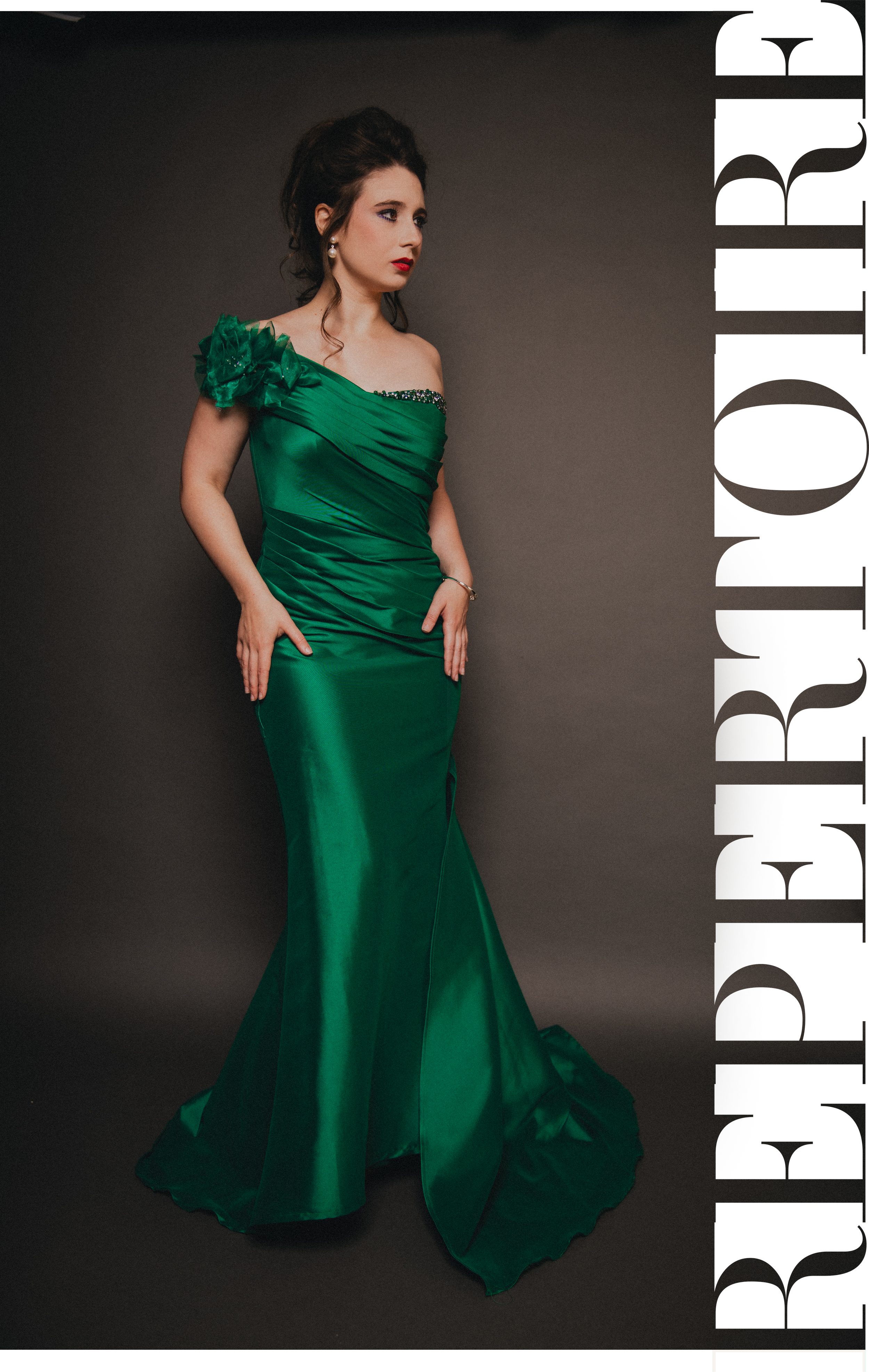 A woman in a long, elegant green satin gown with ruffled shoulder detail, posing against a dark gray background.