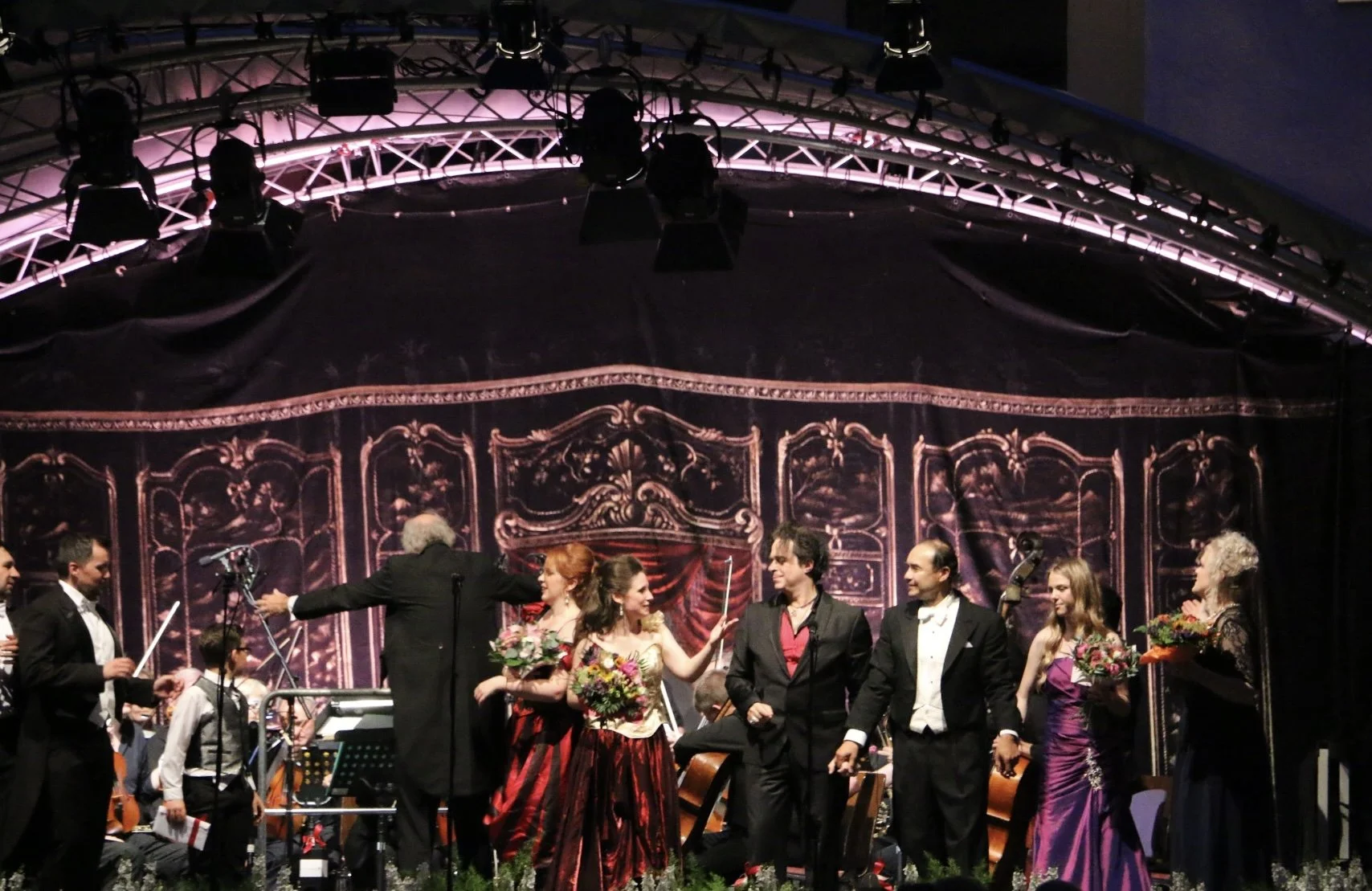 A group of performers on a stage after a concert, bowing and accepting flowers, with an orchestra in the background and a decorated curtain backdrop.