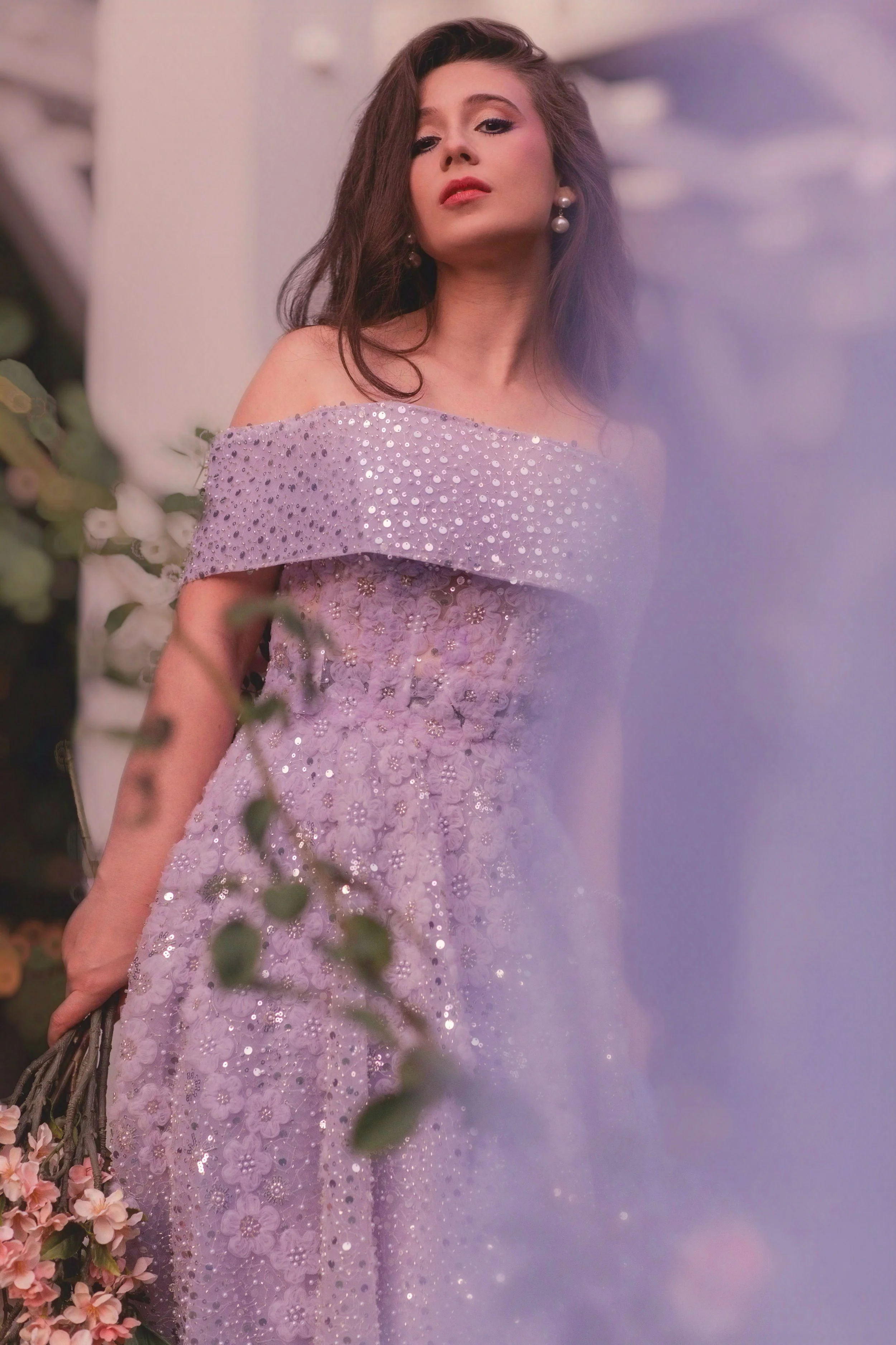 A woman in an off-the-shoulder lavender gown with sparkling embellishments, holding a bouquet of pink flowers, standing outdoors near white columns.