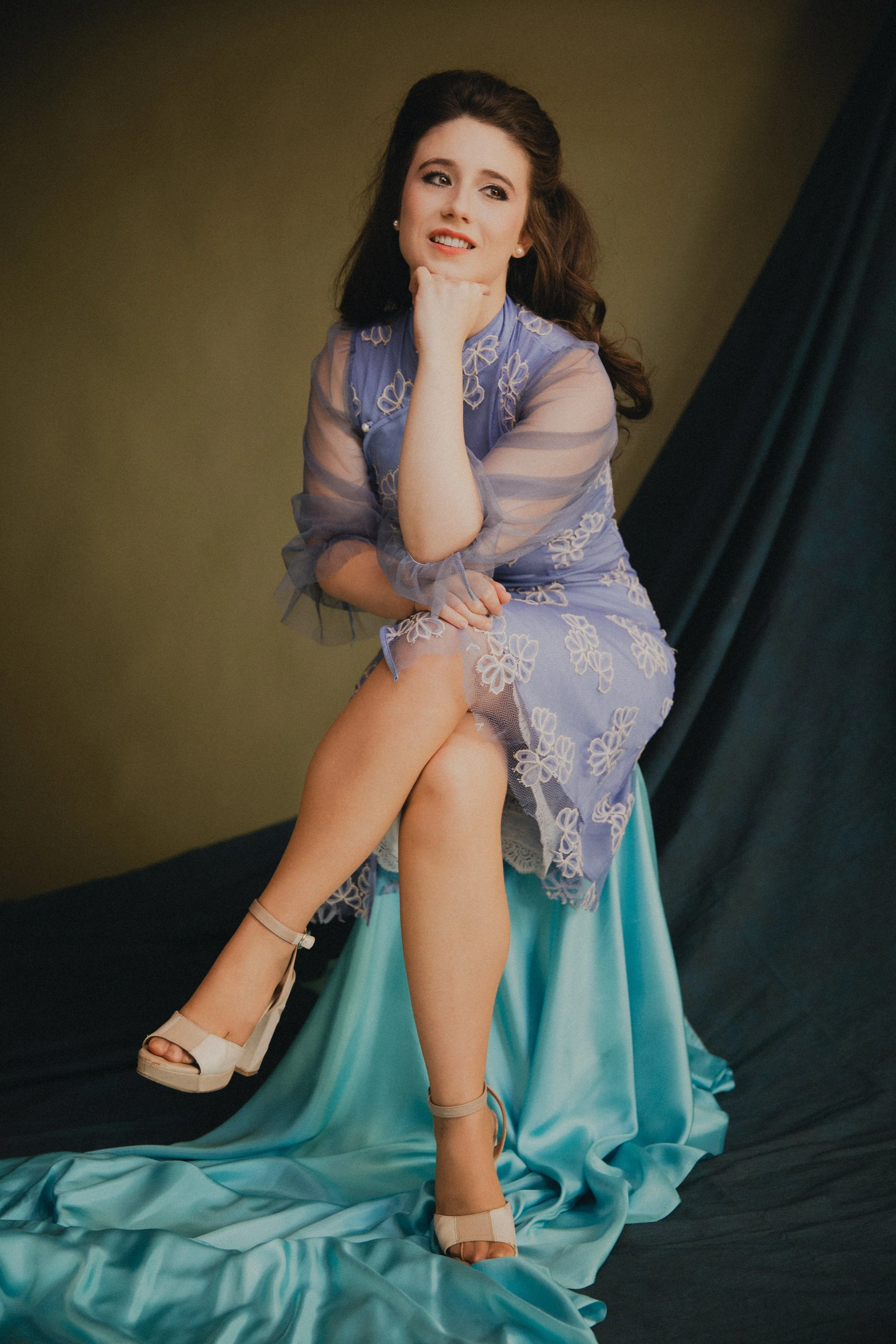A woman sitting on a teal fabric-covered stool, wearing a sheer purple dress with floral embroidery and high heel shoes, posed with her chin resting on her hand and crossed legs, against a plain background with a dark curtain.