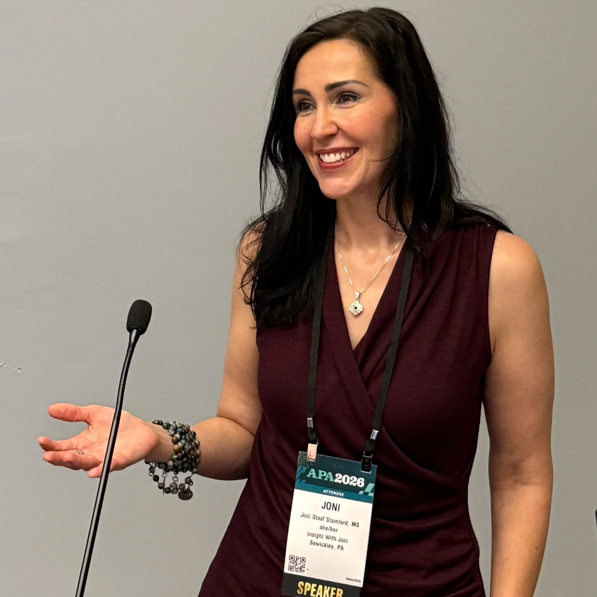 A woman with long dark hair smiling, wearing a maroon sleeveless top and multiple beaded bracelets, standing at a podium with a microphone. She has a conference badge that reads "Joni," with additional text indicating her participation in a speaking event at an APA 2026 conference. The background is a plain gray wall.