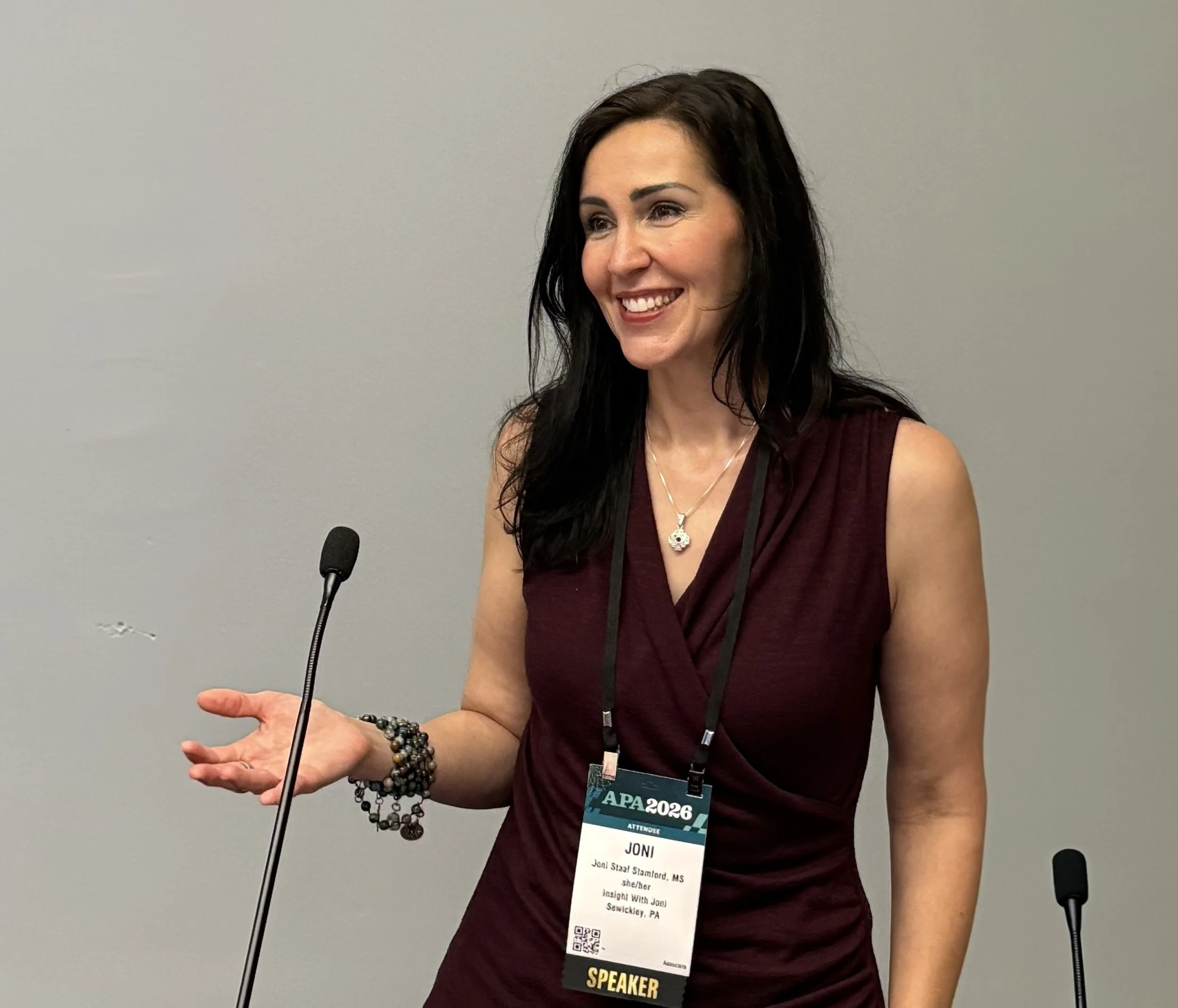 A woman with long dark hair smiling and speaking at a conference, wearing a maroon sleeveless top, multiple beaded bracelets, a necklace, and a conference badge that reads "Joni" and "Speaker".