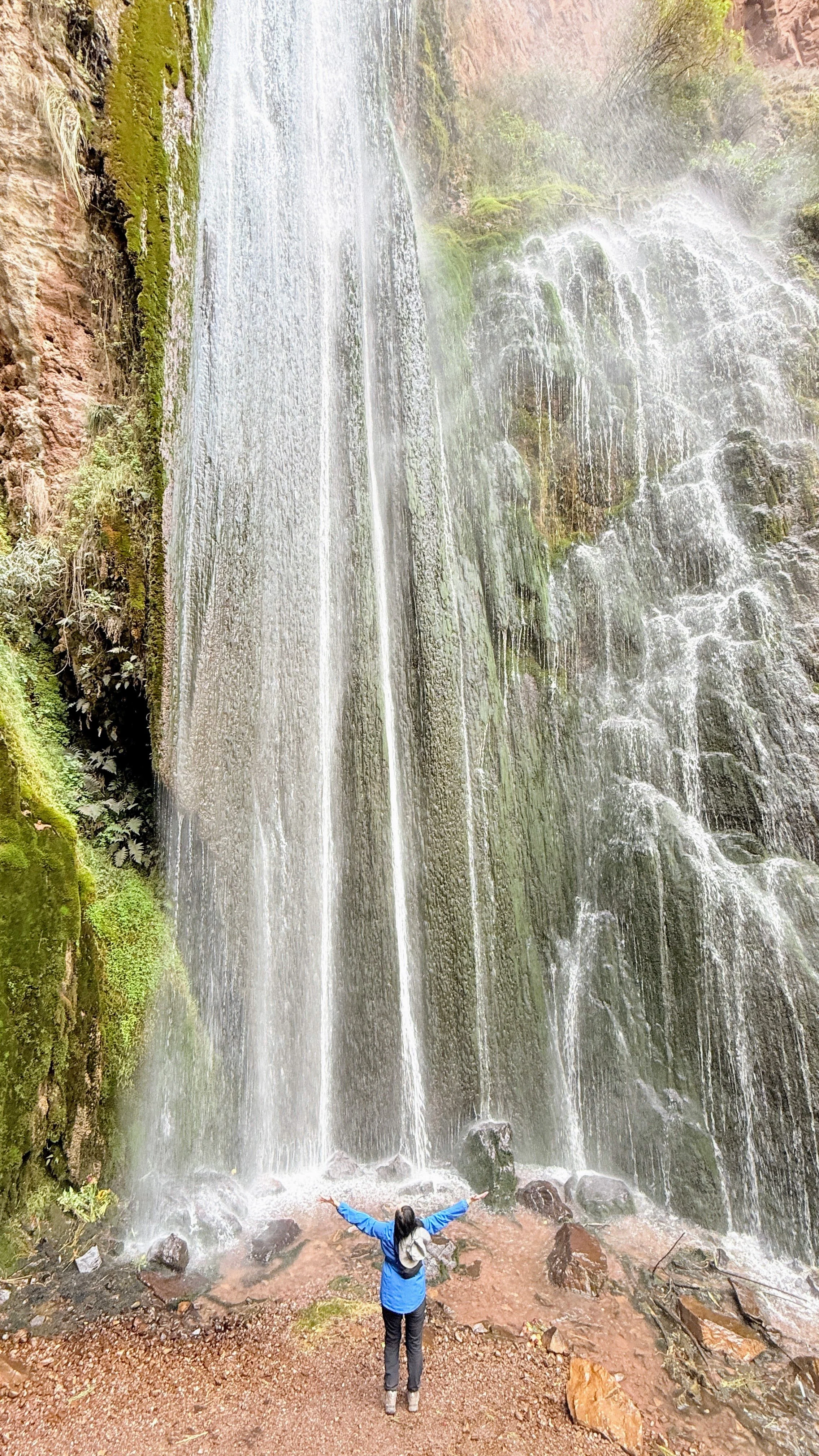 Joni Staaf-Stamford standing with arms outstretched in front of a tall waterfall in Peru's Sacred Valley.