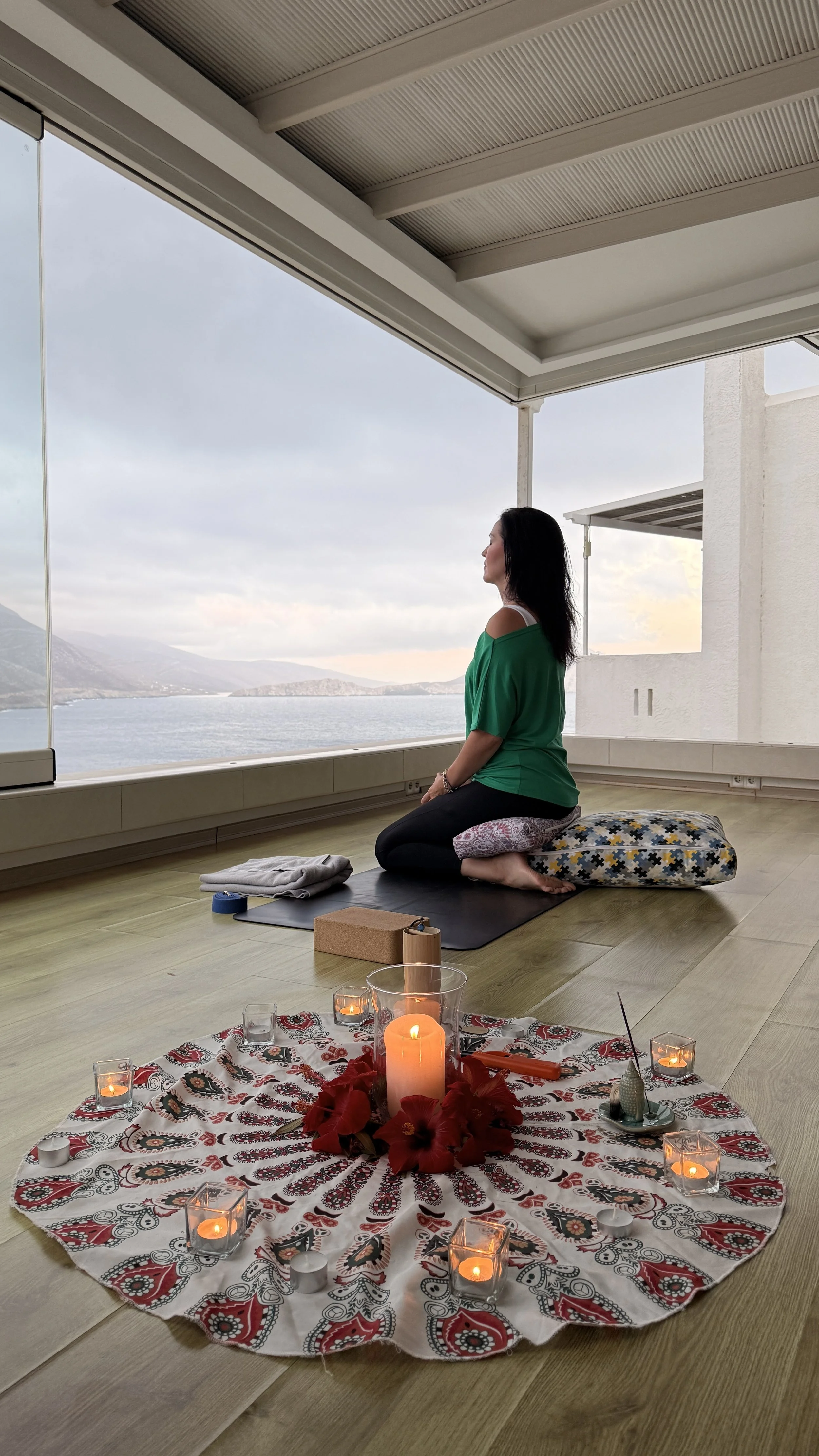 A woman practicing meditation in a peaceful room with a scenic water view. The room has a low table decorated with candles, flowers, and a patterned cloth.