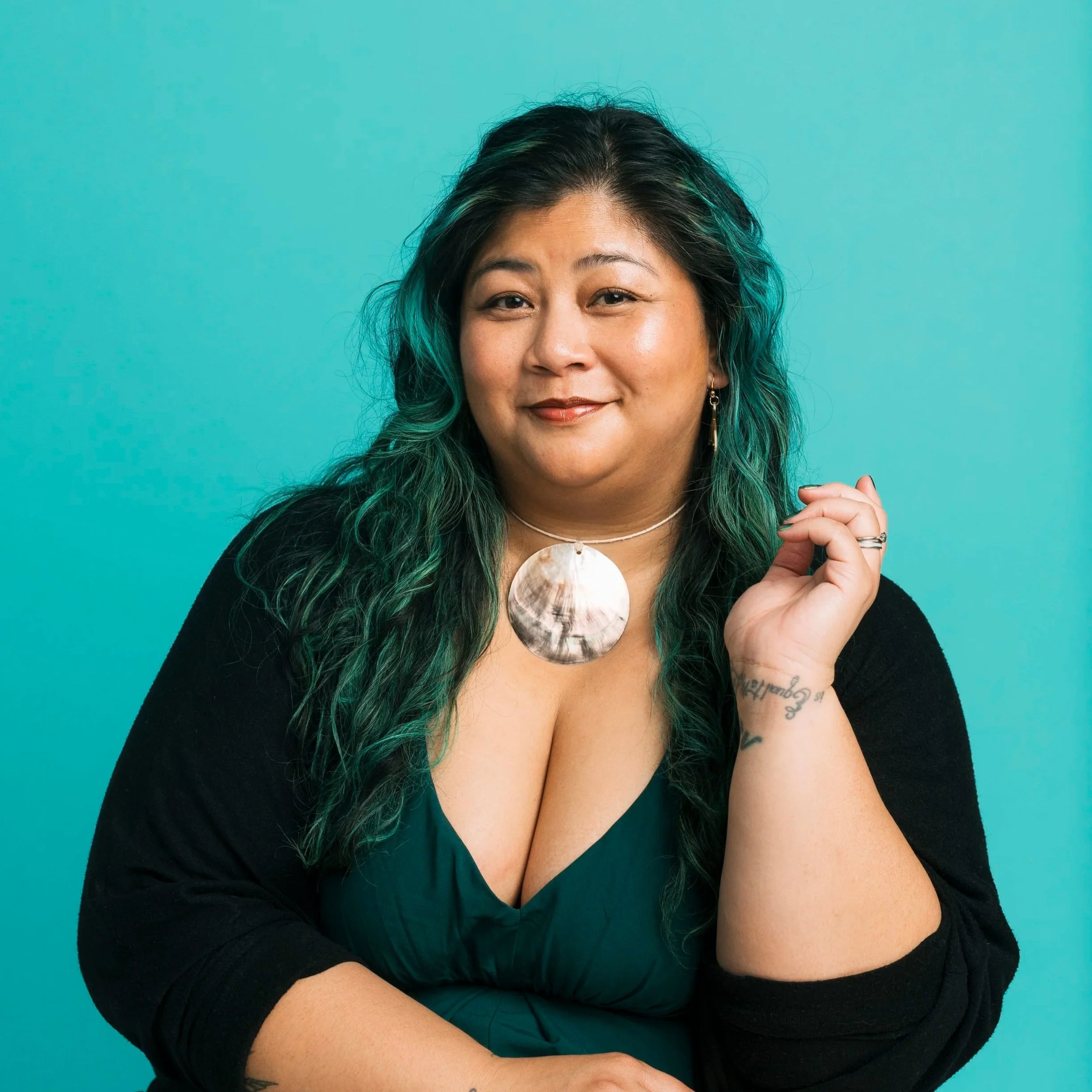 A Filipina with black and green hair, looking at the camera with a slight smile. She is wearing a seashell necklace, and a lowcut green drees with a black cardigan. Her left hand is raised and relaxed near her hair.