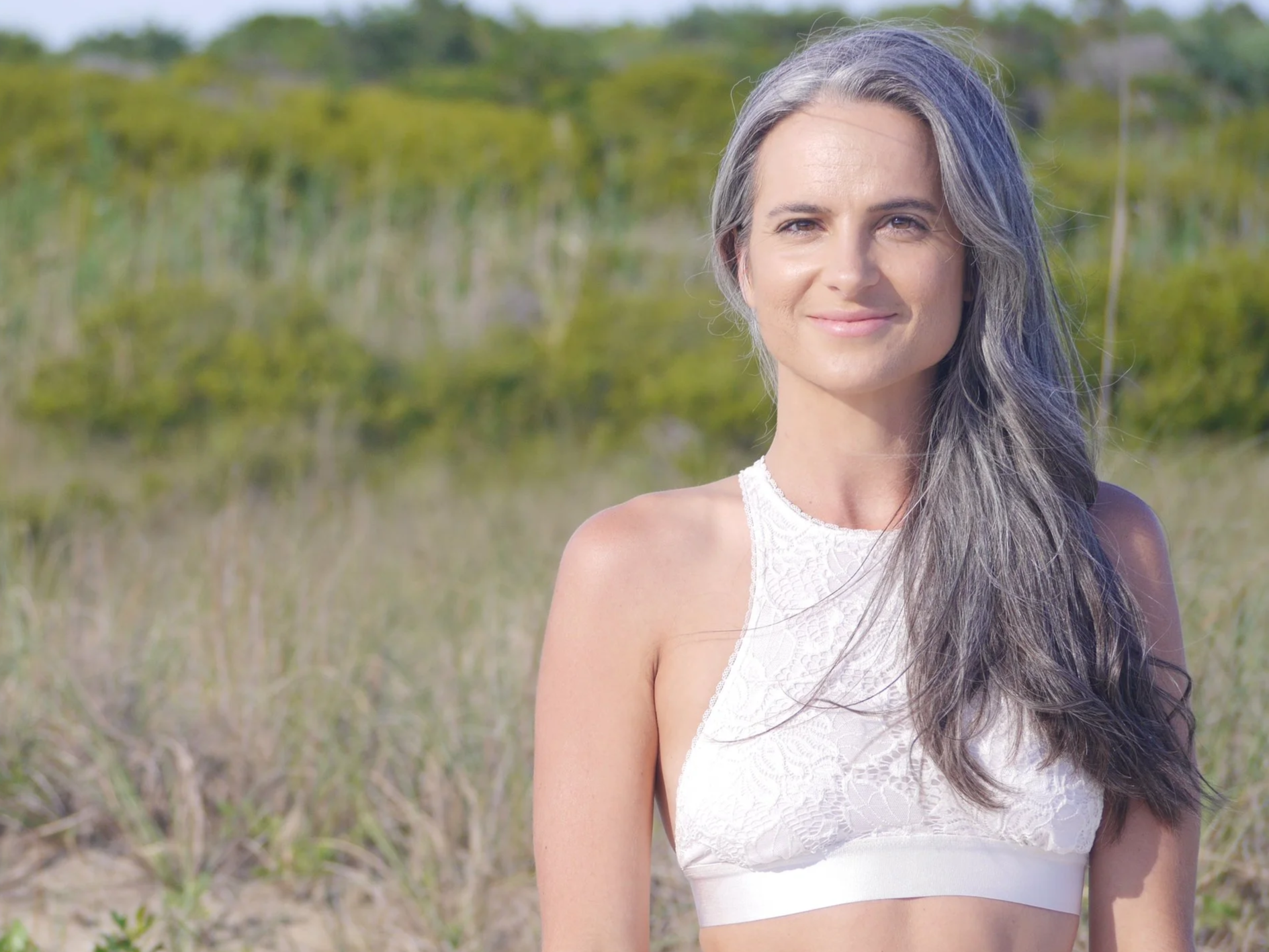 Holistic Health Coach and Ayurvedic Health Counselor, specializing in women's health, Katie Shaw with long gray hair standing outdoors in a grassy area with green trees in the background, wearing a white sleeveless lace crop top.