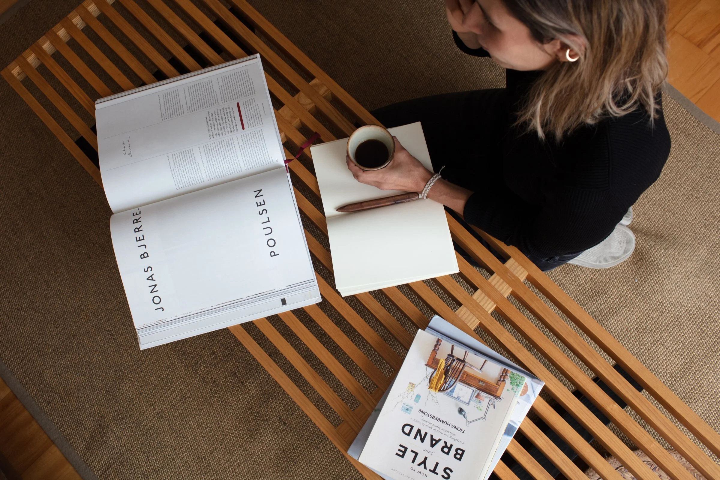 A woman sitting cross-legged on a wooden bench with a cup of coffee, looking at a magazine and a white notebook. There is another magazine titled 'Style Brand' on the bench. An open magazine with the title 'Jonas Buerre' is also visible on the bench.