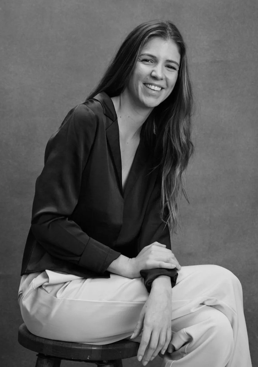 A woman with long hair sitting on a stool, smiling, wearing a dark blouse and light-colored pants, in a posed black-and-white portrait against a plain background.
