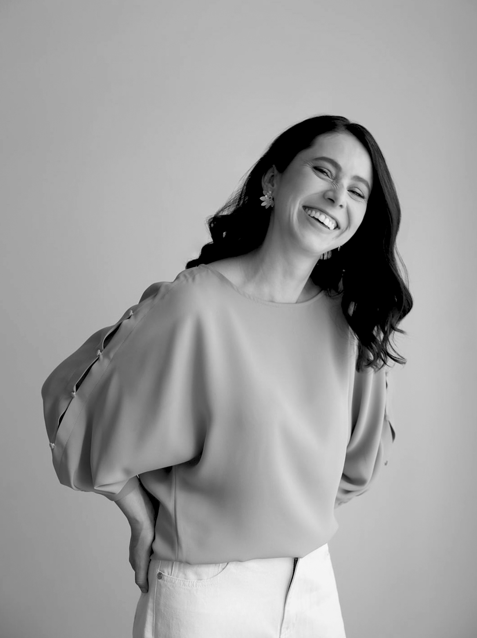 Black and white photo of a woman smiling, wearing a loose top with puffy sleeves and earrings, standing against a plain background.