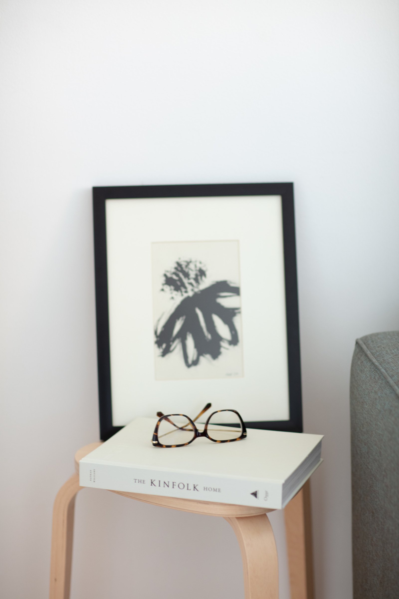 A small light wooden table with a white book titled 'The Kinfolk Home' and a pair of tortoise shell eyeglasses on top. Behind the table, a large black picture frame leans against a white wall, containing an abstract black and white painting or print. To the right, part of a gray upholstered piece of furniture is visible.