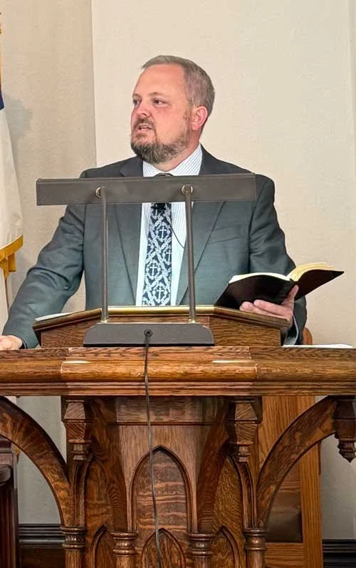 A man standing at a wooden podium in a suit, holding an open book, with a microphone in front of him, in a formal setting.