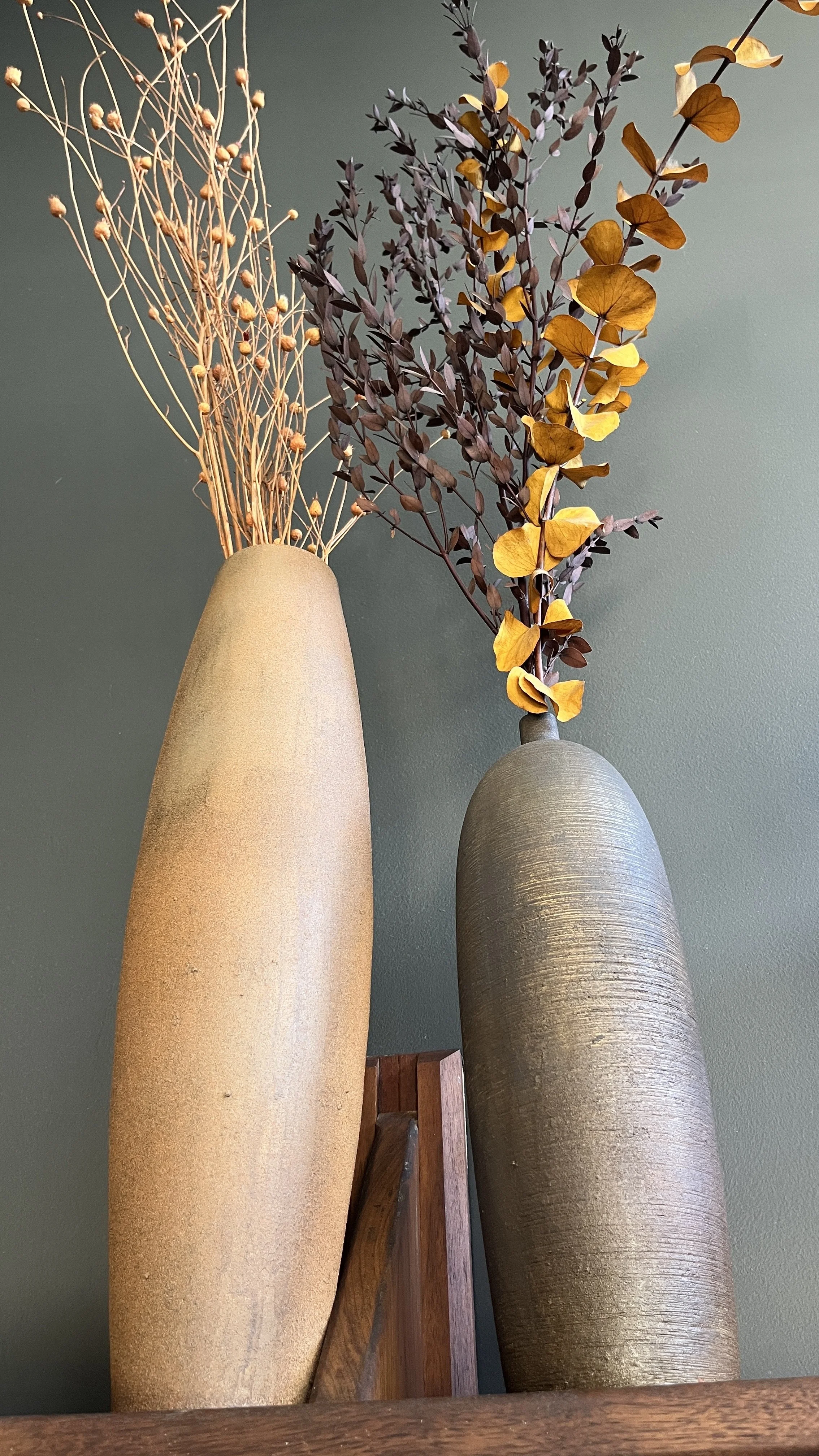 Two tall vases with dried branches and leaves, set against a green wall.