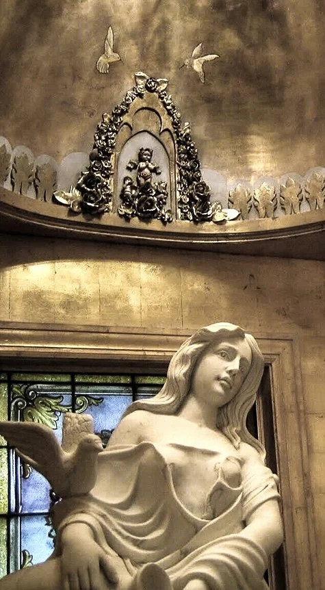 A marble statue of the Virgin Mary with a dove on her arm in a church, with gold accents and religious artwork and imagery in the background.