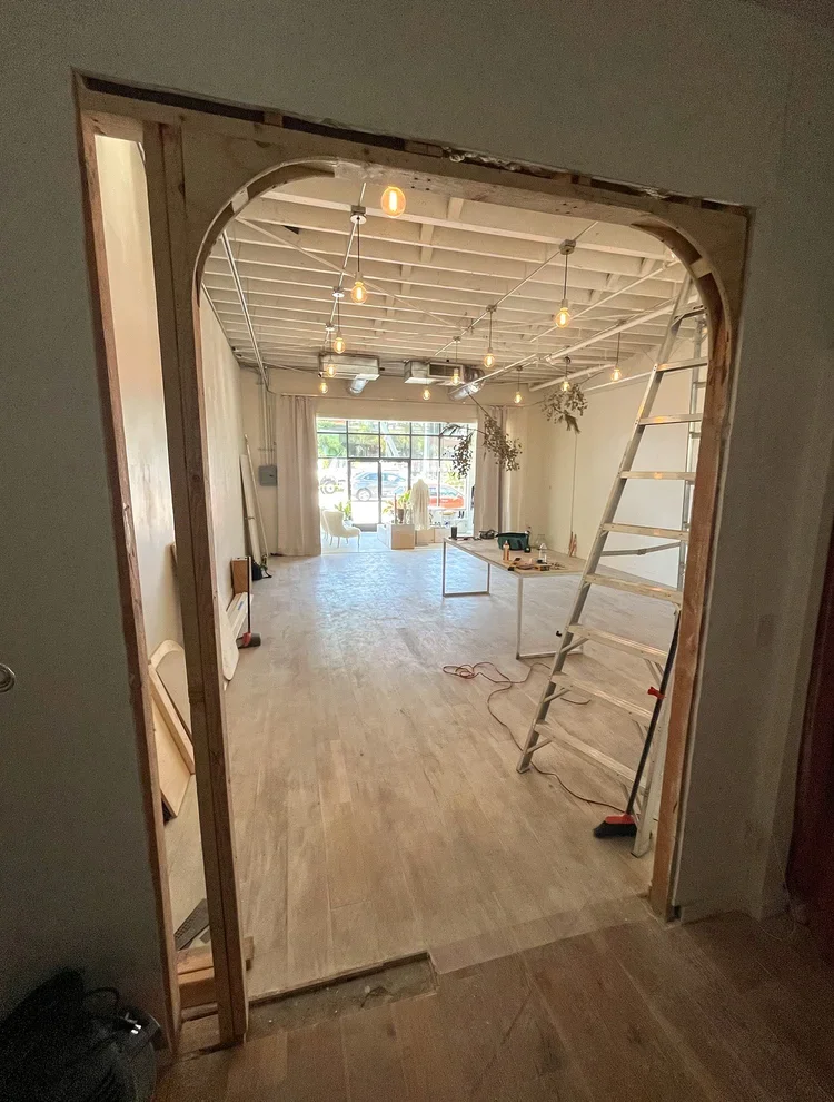 Room under renovation with exposed ceiling beams, string lights, potted plants hanging from ceiling, ladder, work table with tools, large window with curtains, and seating area in the background.