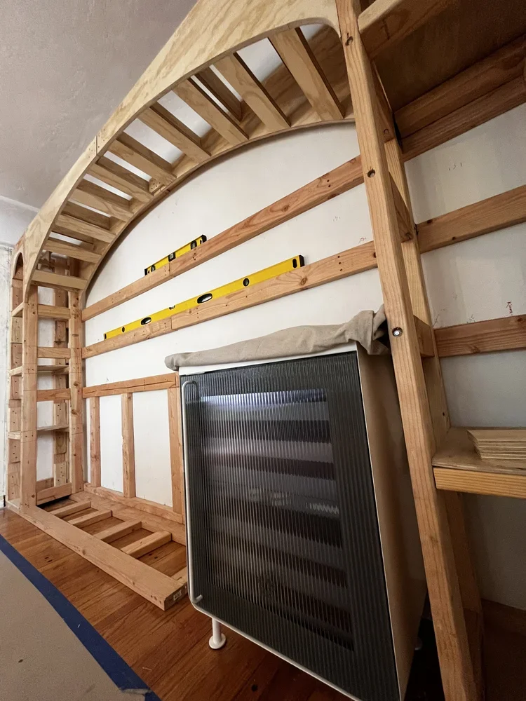 A wooden loft bed frame under construction, with stairs on the left side, a yellow level installed on the wall, and a black space heater in front.