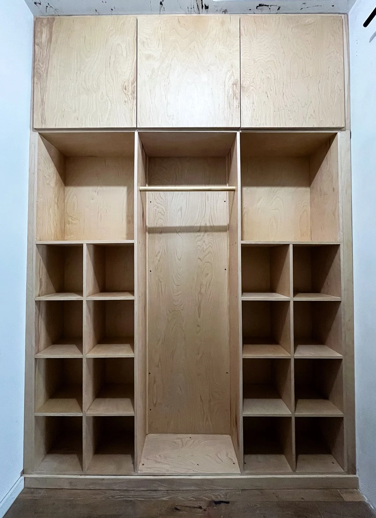 Unfinished wooden closet with multiple compartments, open shelves, and a hanging rod in the middle, against a white wall.