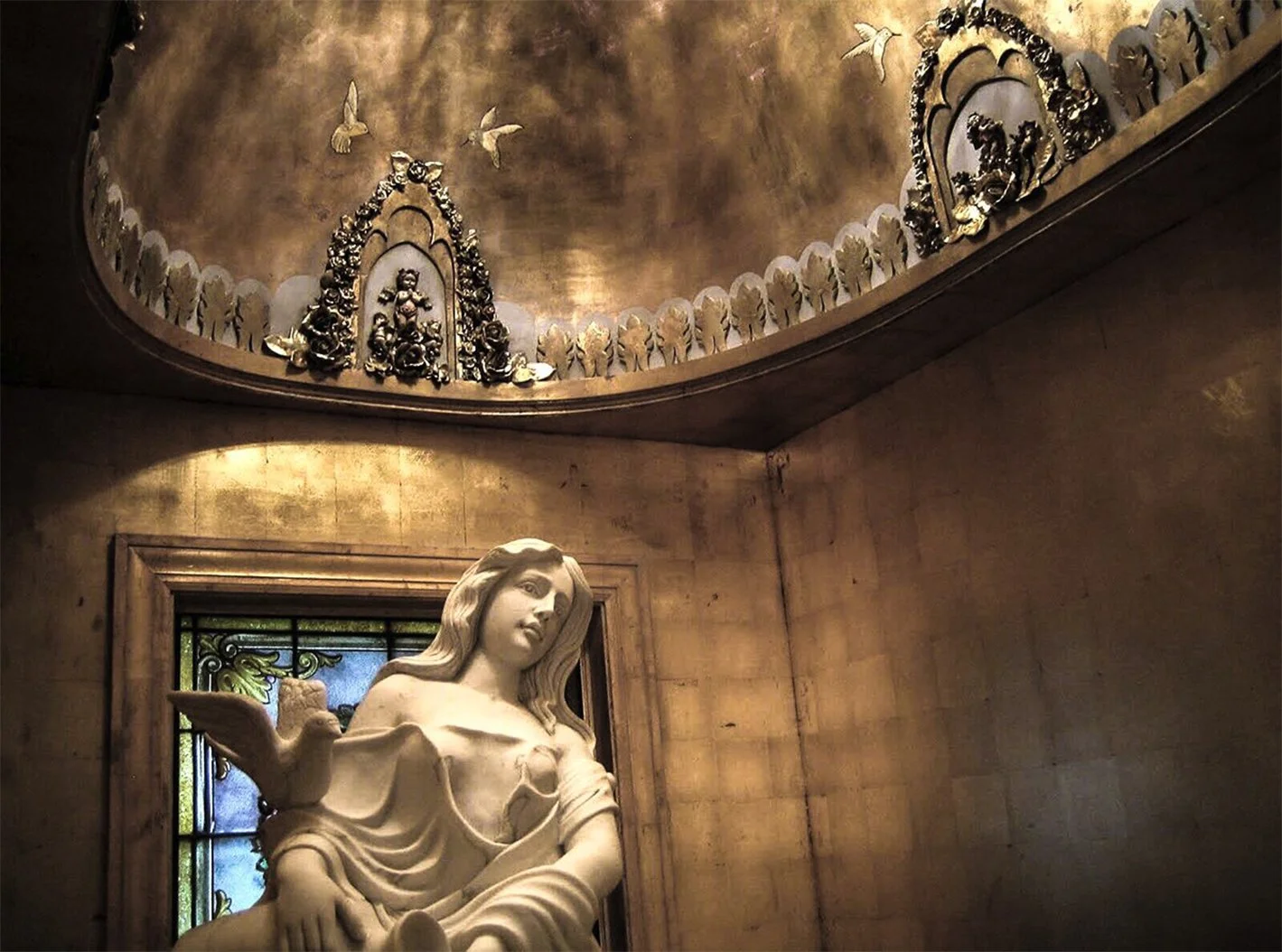Interior of a church featuring a white statue of a woman with a dove on her shoulder, stained glass windows behind her, and ornate gold decorations on the ceiling with religious figures and birds.