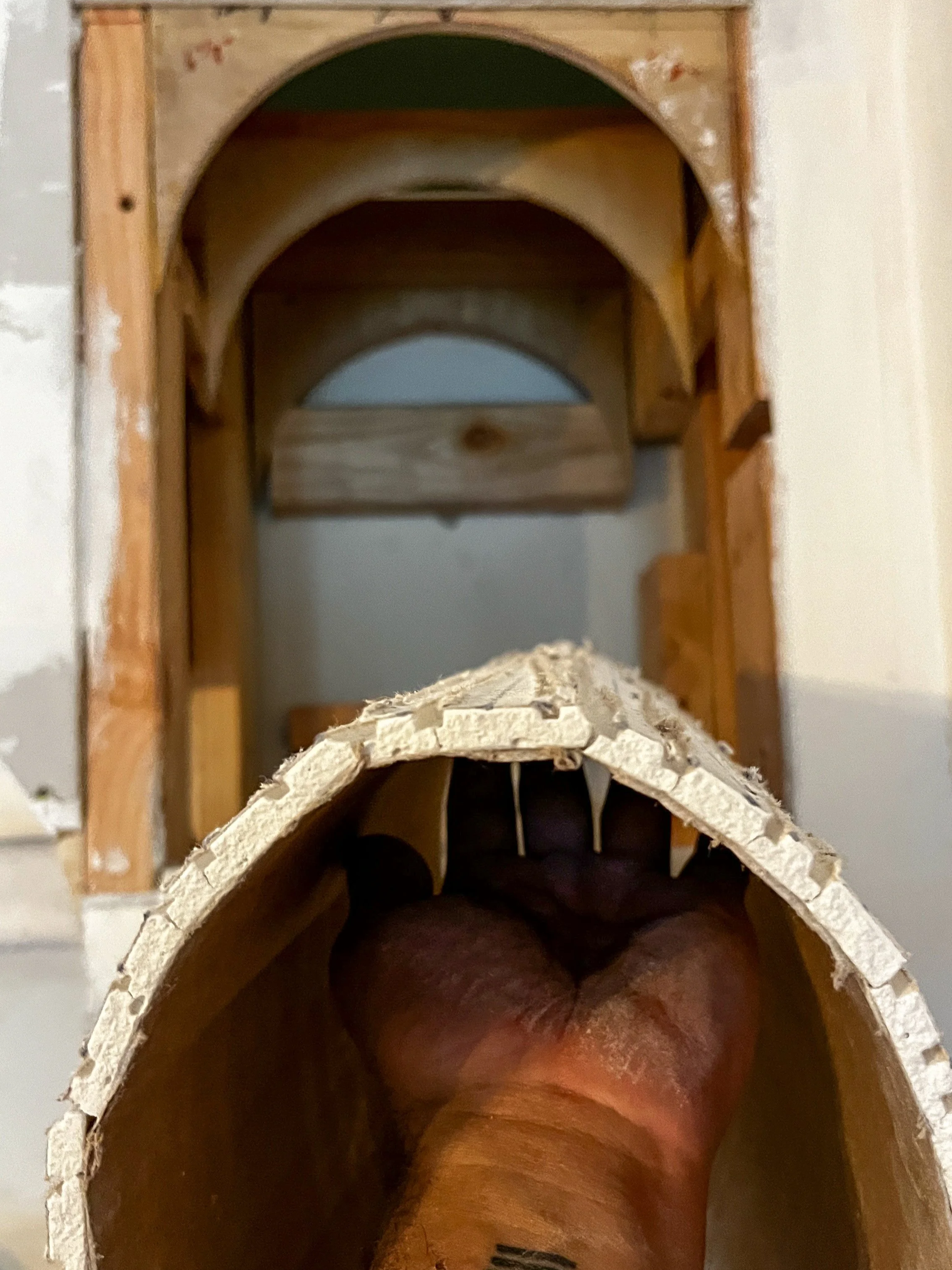Close-up of a person's hand holding a section of a church organ pipe with an unfinished wooden structure in the background.