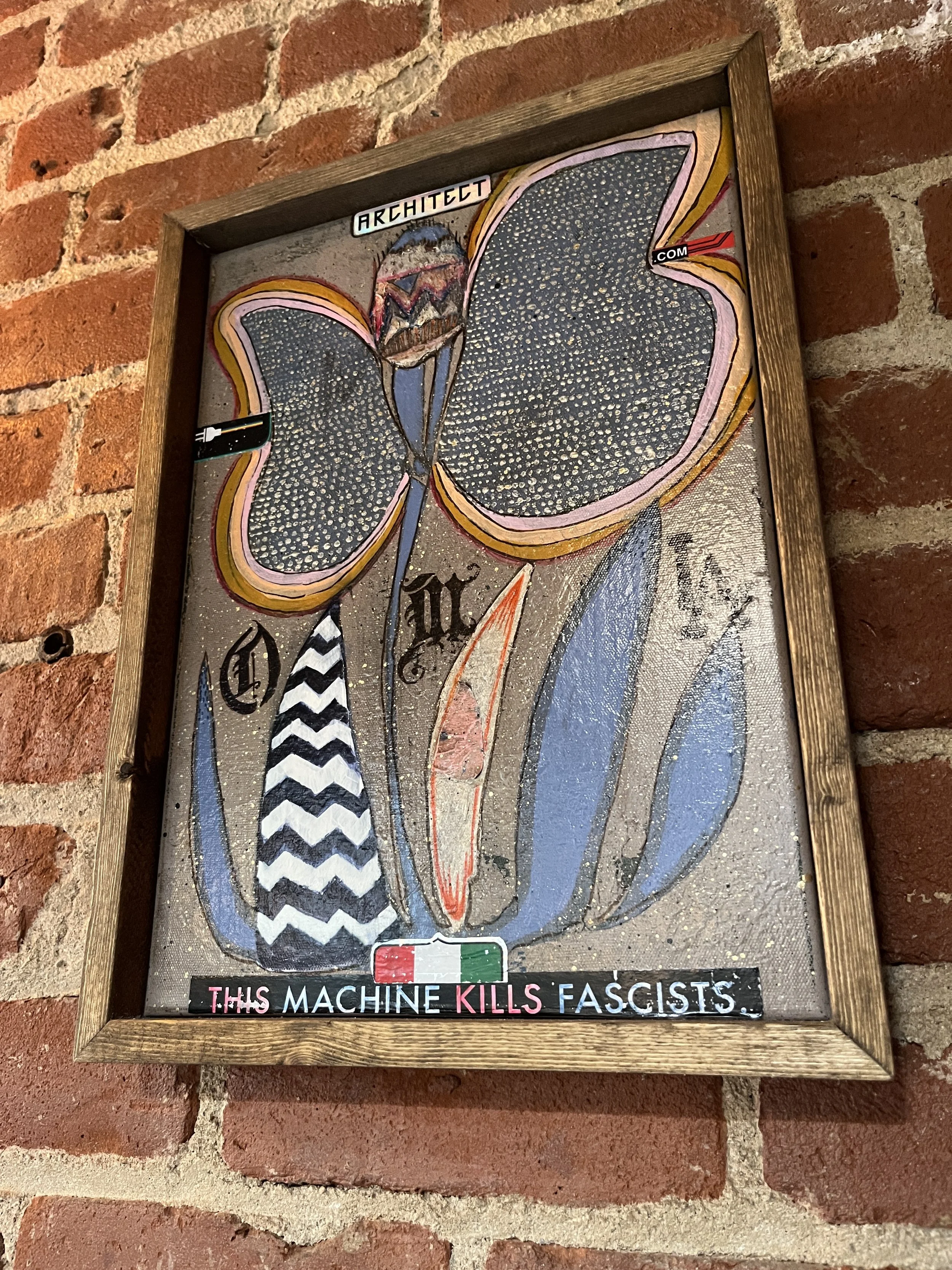 Artwork of a butterfly with blue and pink wings, with text on the framed artwork saying 'ARCHITECT.COM' at the top and 'THIS MACHINE KILLS FASCISTS' at the bottom, against a red brick wall custom painting bar hospitality wooden frame.