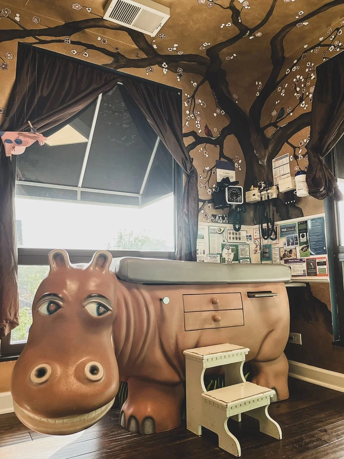 Decorated room with a mural of a cherry blossom tree on the ceiling, a window with dark curtains, and a hippopotamus-shaped bed with a smiling face and eyes. There is a small white step stool in front of the bed, and a bulletin board with notices and equipment on the wall.