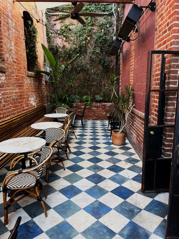 An outdoor patio with checkered black and white tile floor, seating area with marble-topped tables and rattan chairs with black and white patterned cushions, brick walls, string lights, and green plants along the back wall.