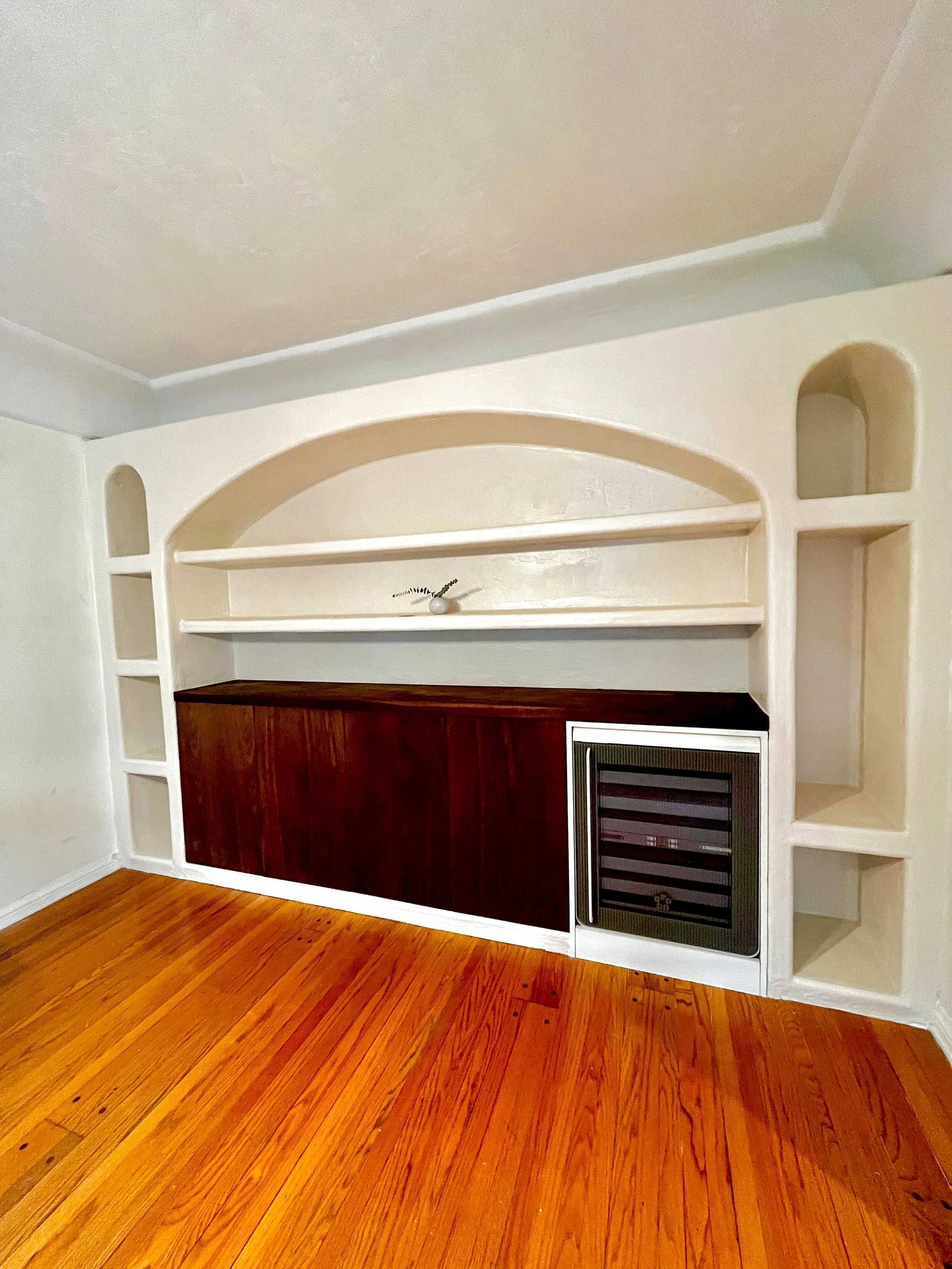 Living room with a built-in white bookshelf, dark wood cabinet, and small black refrigerator on a hardwood floor.