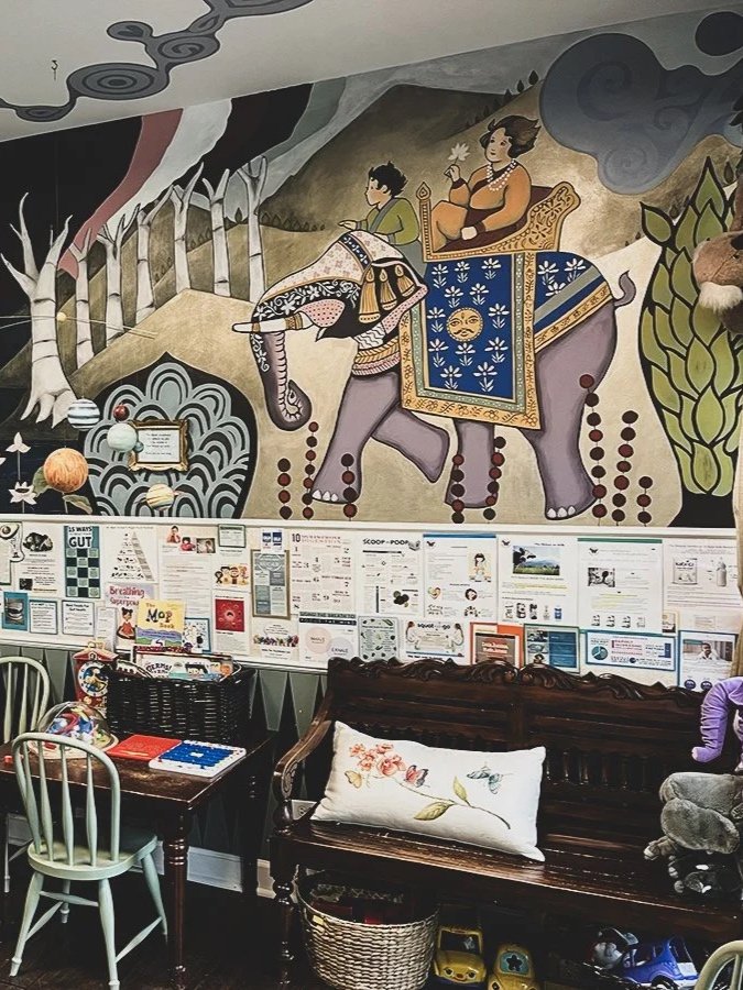 Decorative mural of an elephant carrying two women, one reading a book and the other holding incense, surrounded by plants and landscape, with children's toys and papers on a table and a wooden bench with a floral pillow below.