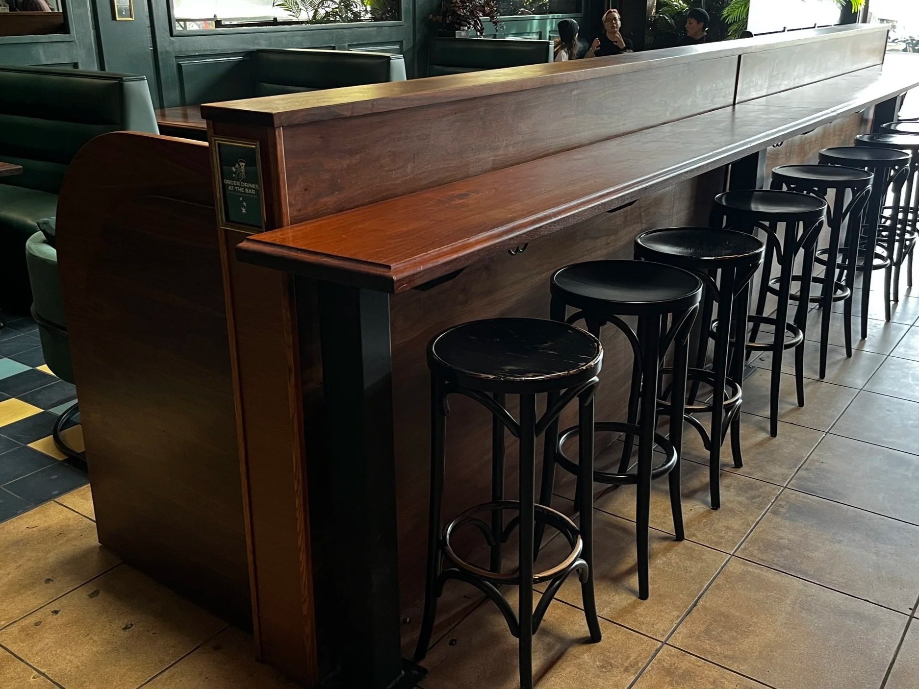 A row of black bar stools lined up in front of a high wooden bar counter in a restaurant or cafe with tiled flooring and some green chairs and people in the background.