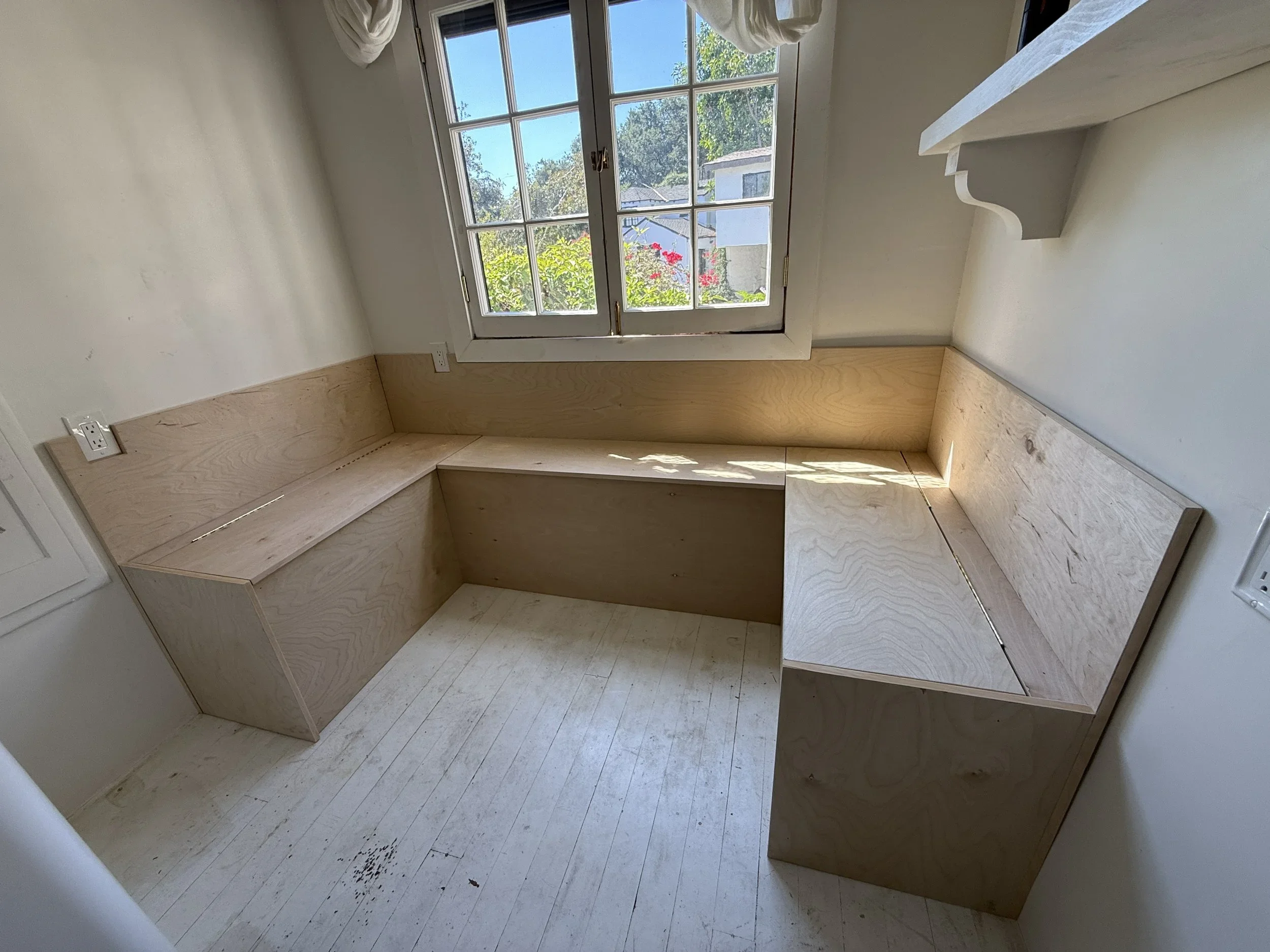 Empty room with a large window, built-in wooden bench seating along two walls, and a shelf on the wall.