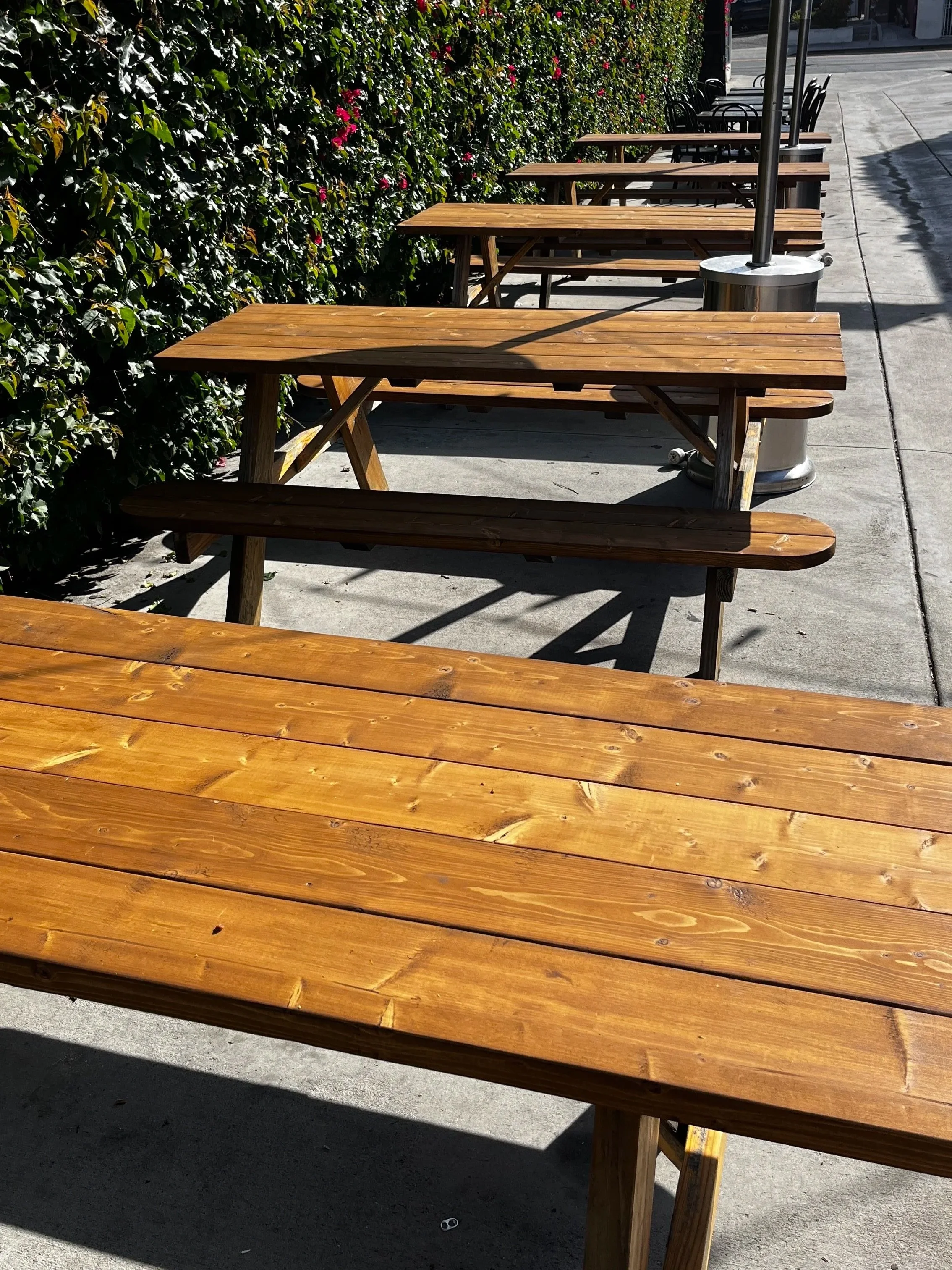 Multiple outdoor wooden tables and benches lined up along a sidewalk with a hedge and pink flowers in the background, and a large patio umbrella and a trash can nearby.