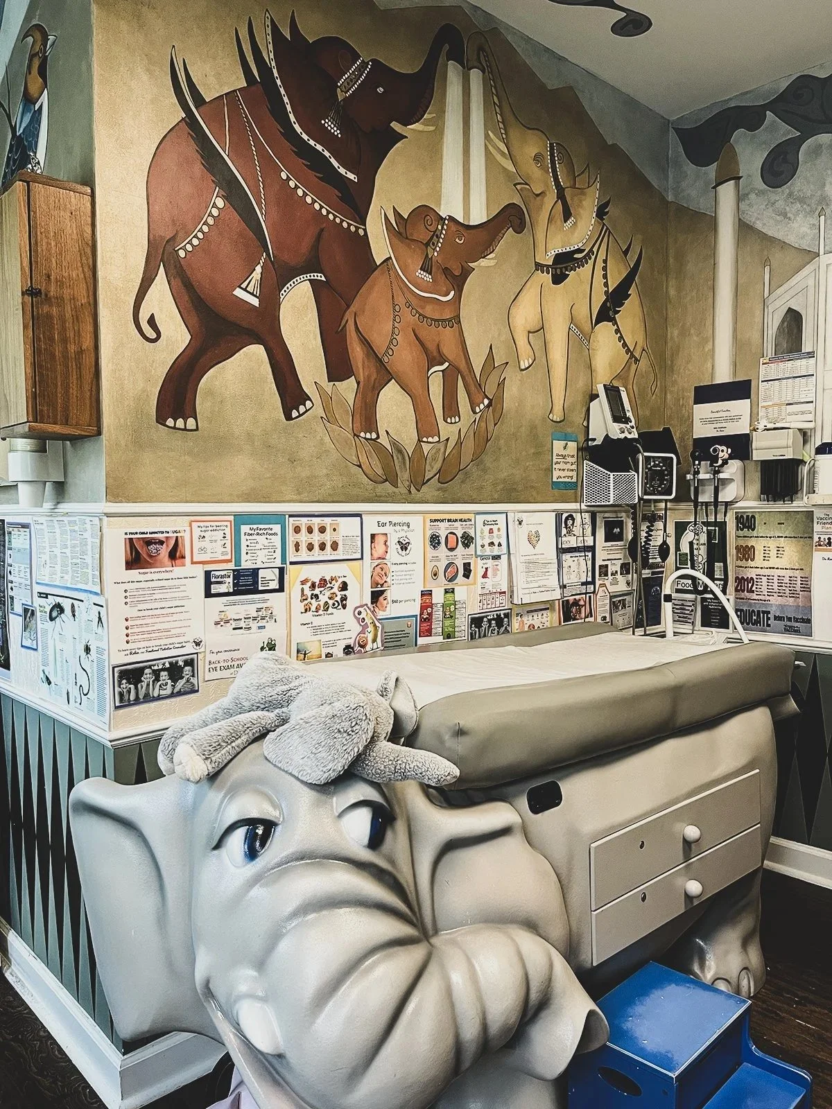 Hospital room or clinic with a mural of elephants on the wall, medical equipment on the side, informational posters, and a large Elephant-shaped chair or decoration in the foreground.