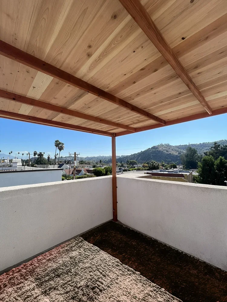 Corner of a balcony with a wooden ceiling and white concrete walls overlooking a cityscape with palm trees, hills, and a clear blue sky.