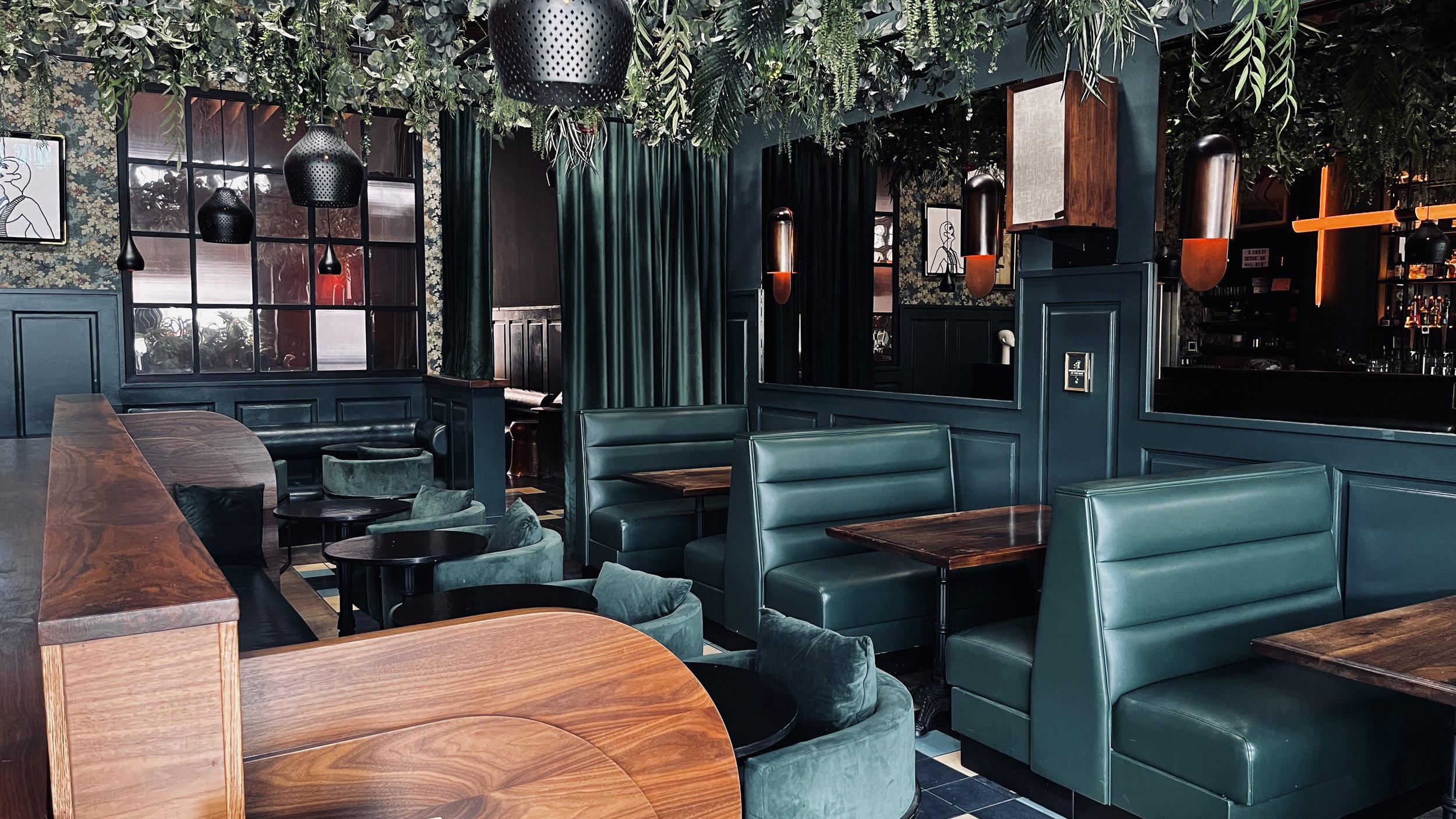 Interior of a modern restaurant or bar with green booths, velvet chairs, dark wood tables, hanging black pendant lights, and greenery on the ceiling.