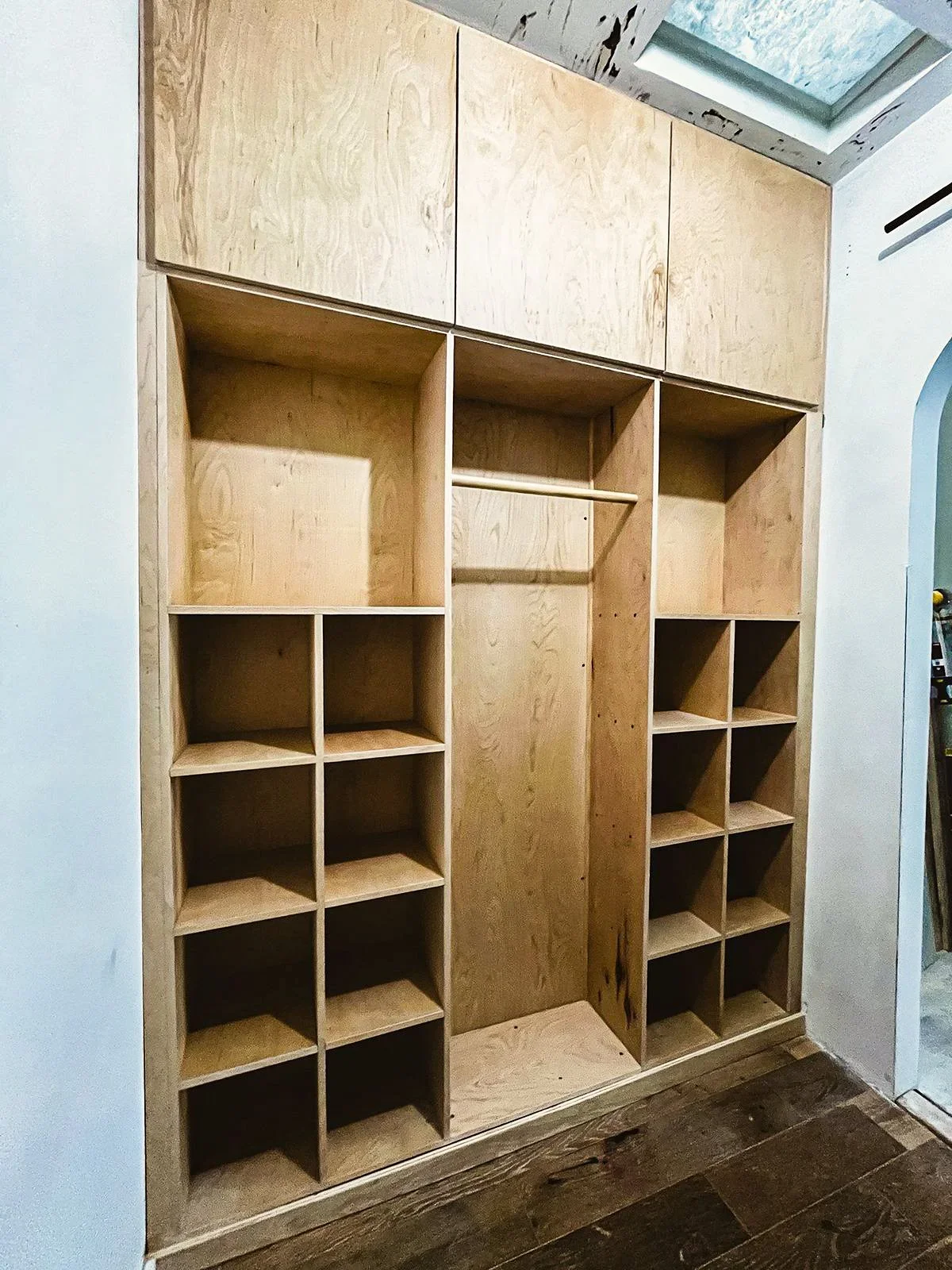 Unfinished wooden closet with multiple cubbies and a hanging rod, installed on a wall with a wooden floor.