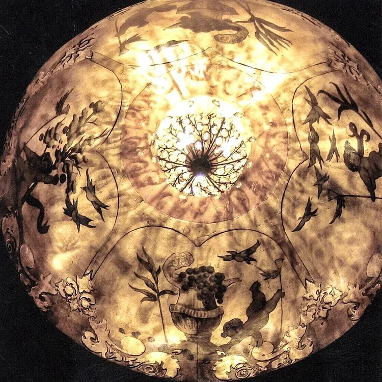 A globe-shaped paper lantern with decorative illustrations of birds, a fruit basket, and ornate patterns. The lantern is lit from within, creating a warm glow.