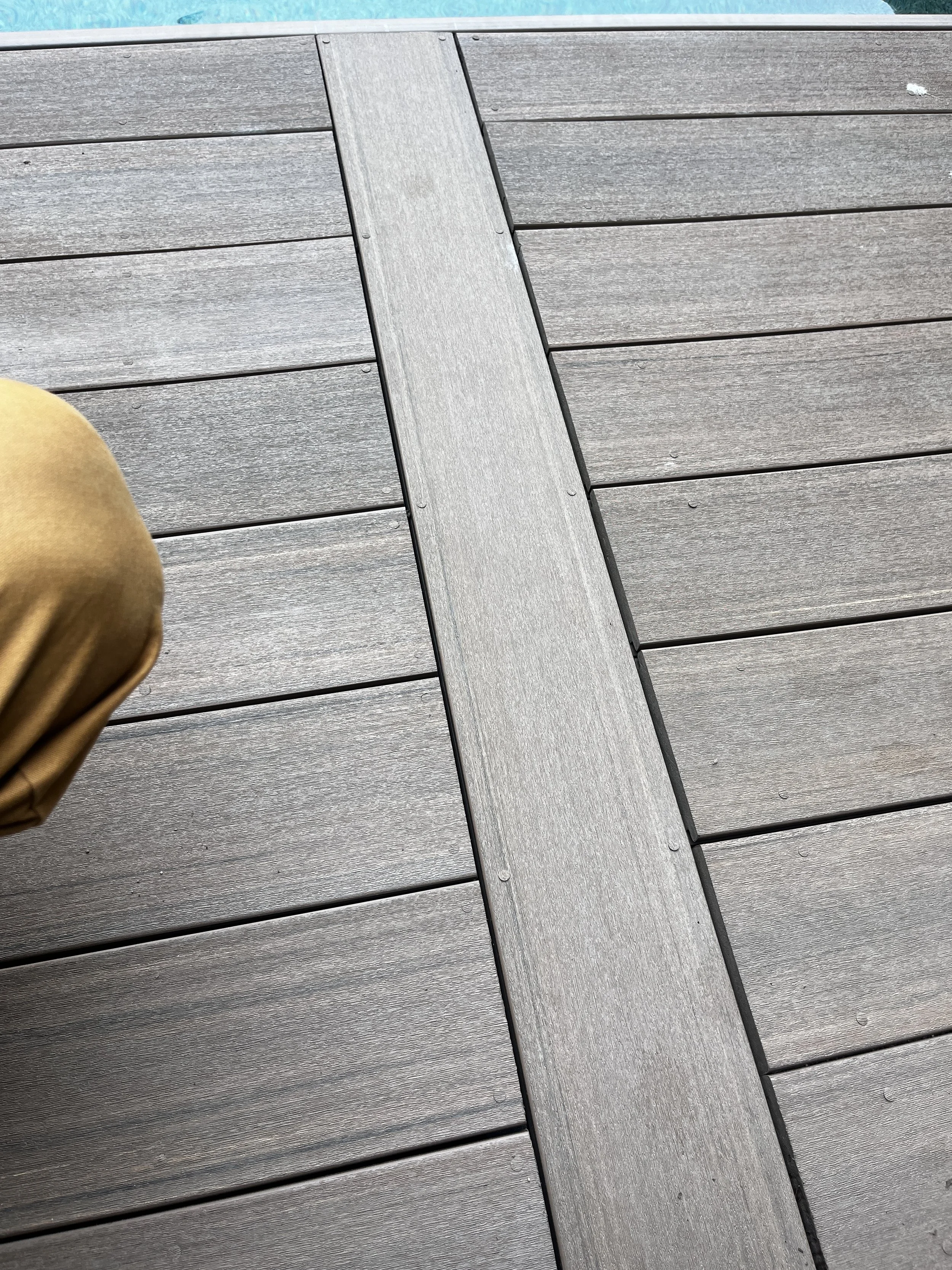 Close-up photo of a wooden outdoor deck with horizontal planks and a central grey-colored strip, with part of a person wearing tan clothing visible in the lower left corner.