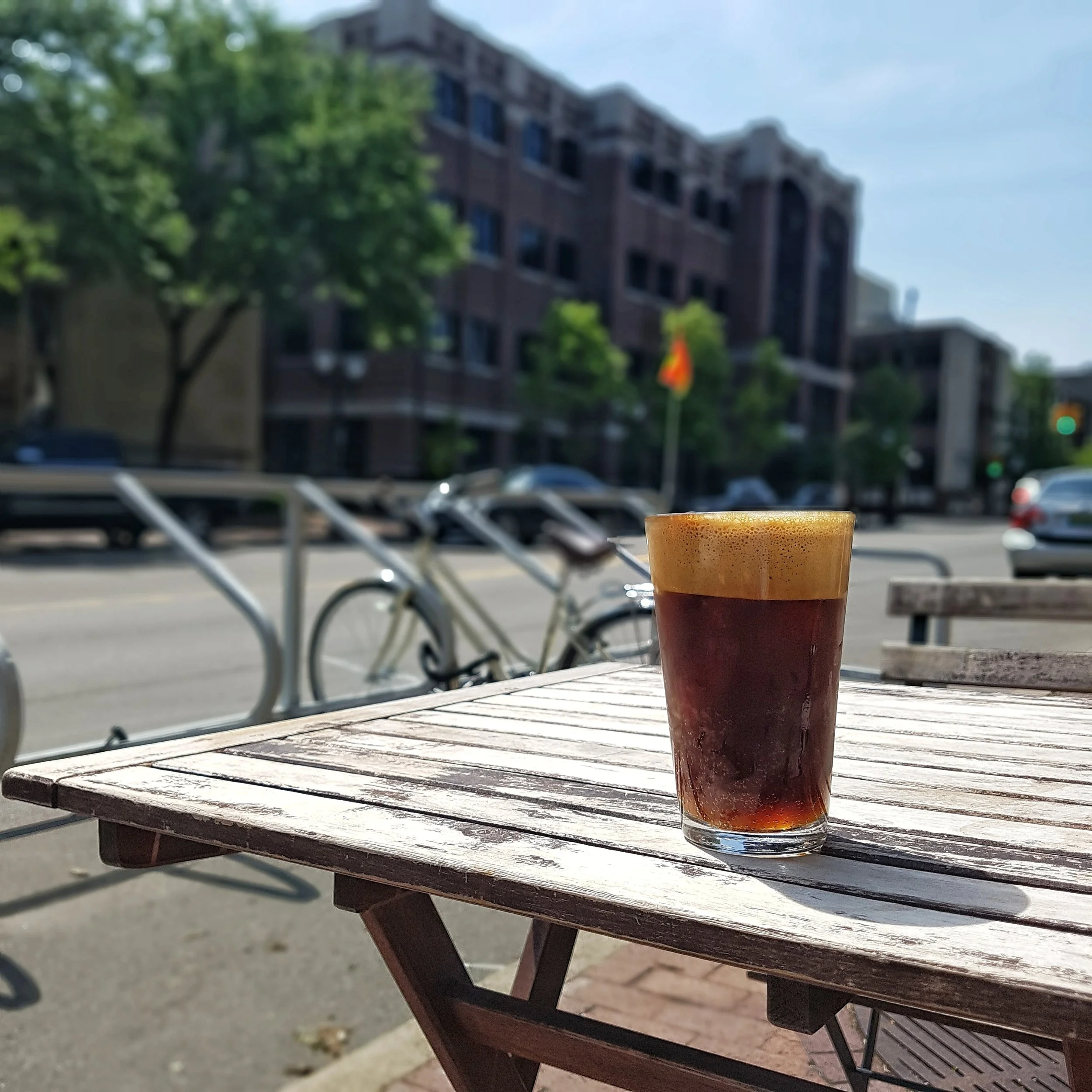 A glass of espresso tonic on a wooden table in the foreground, with a city scene in the background
