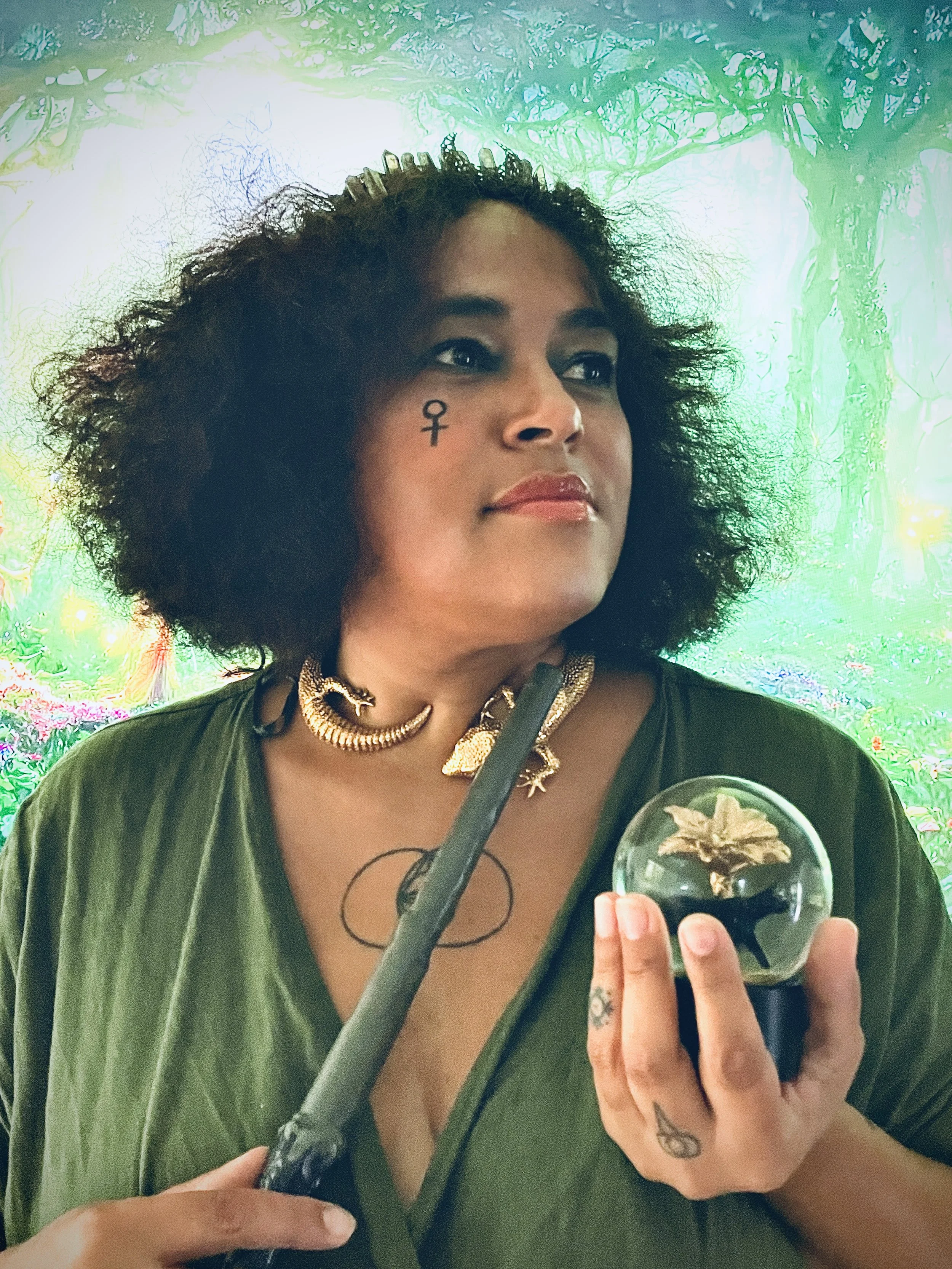 Woman with tattoos on her hand, wearing a choker necklace and a dark green top, holding a crystal ball with a flower inside, in front of a vibrant green forest-themed background.