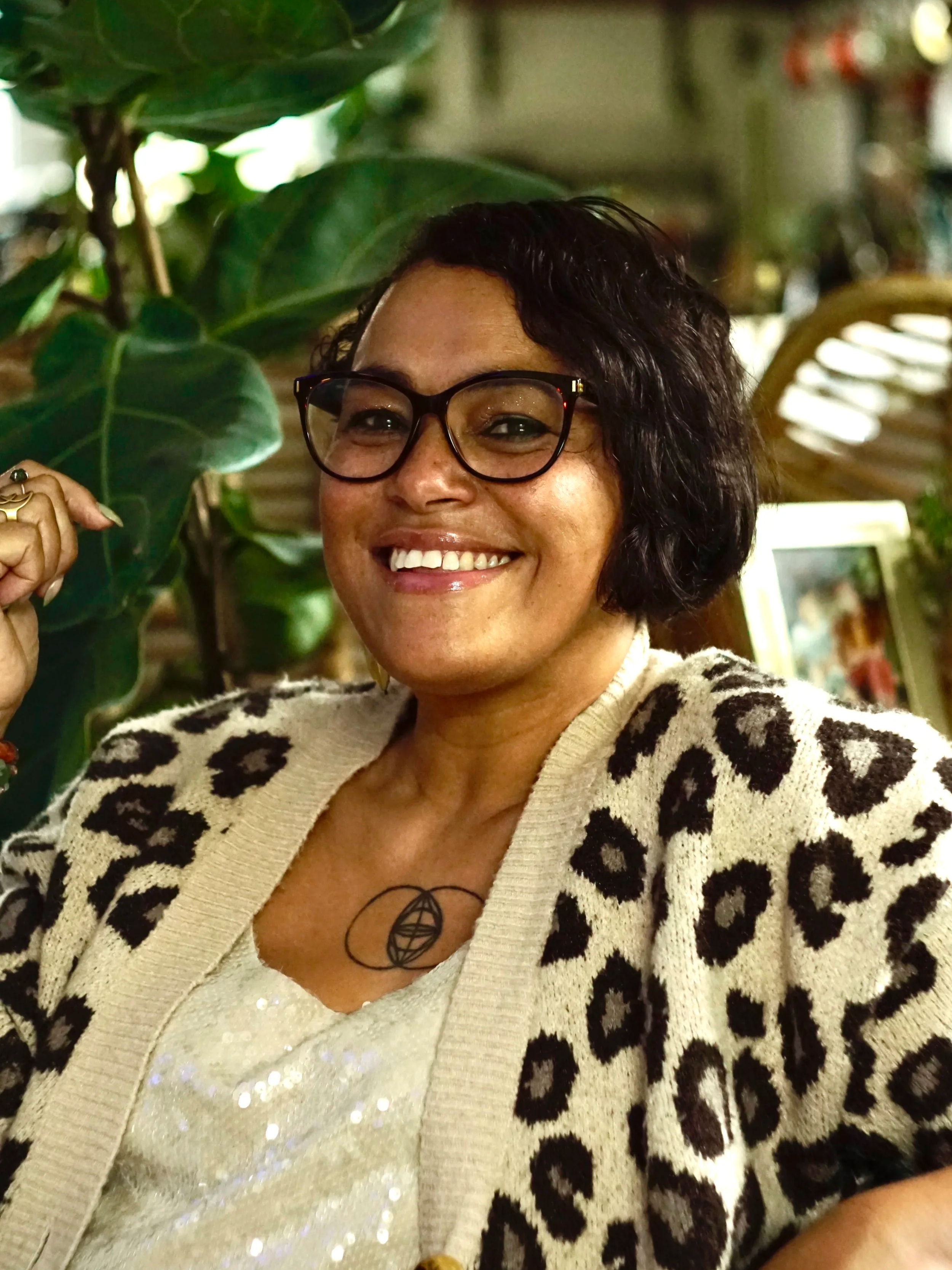 A woman with short curly black hair, glasses, and a wide smile, wearing a leopard print cardigan over a sparkly top, sitting in front of green plants and a mirror.
