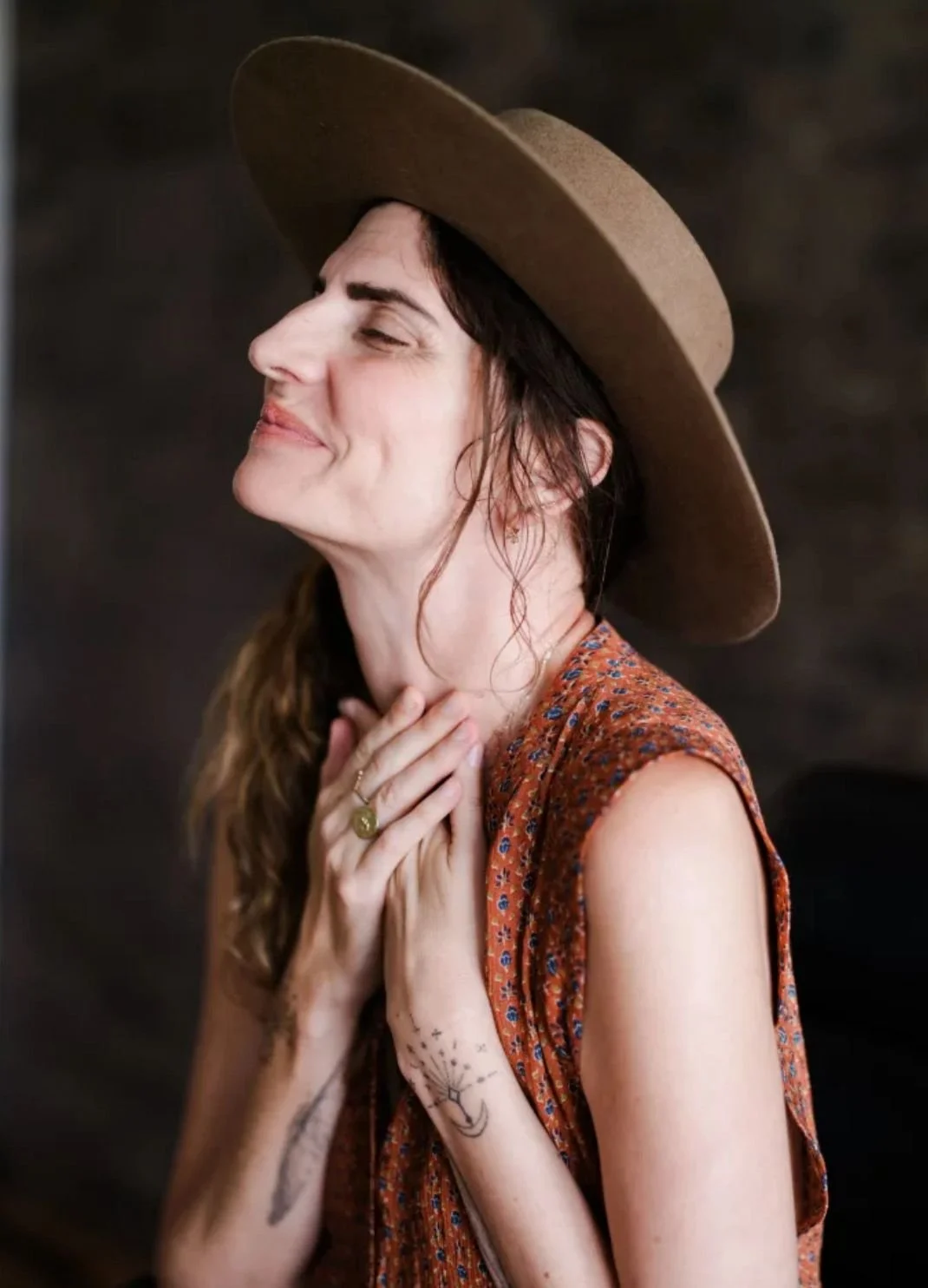A woman with long brown hair wearing a wide-brimmed brown hat and a sleeveless patterned orange dress, smiling softly with her eyes closed, touching her neck and chest, against a dark background.