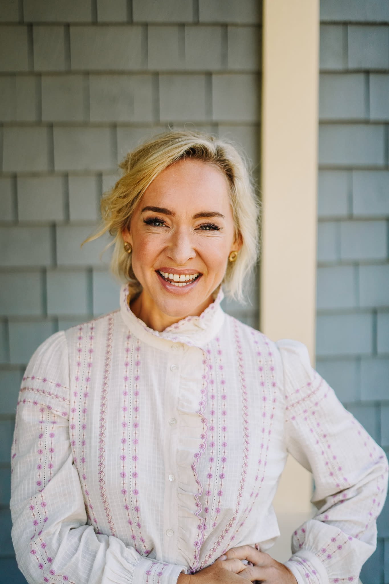 A smiling woman with blonde hair wearing a white blouse with pink embroidery, standing in front of a gray siding wall with a yellow vertical trim.