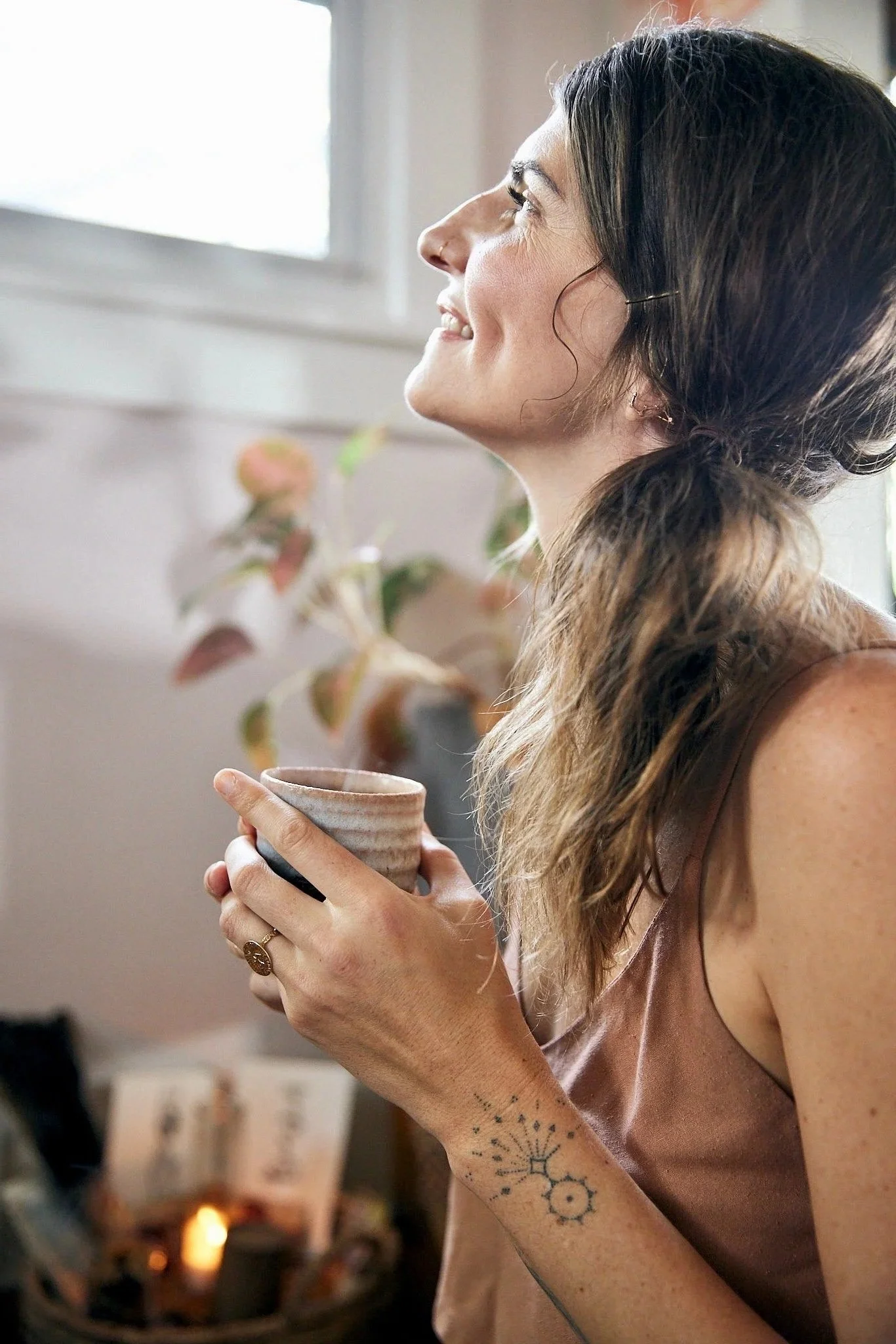 A woman with long curly brown hair, tattoos on her left arm, and wearing earrings, is holding a ceramic mug with both hands and smiling while looking to the right. In the background, there is a fireplace with a burning fire and decorative items on the mantel, including a lantern. The room appears cozy and warmly lit.