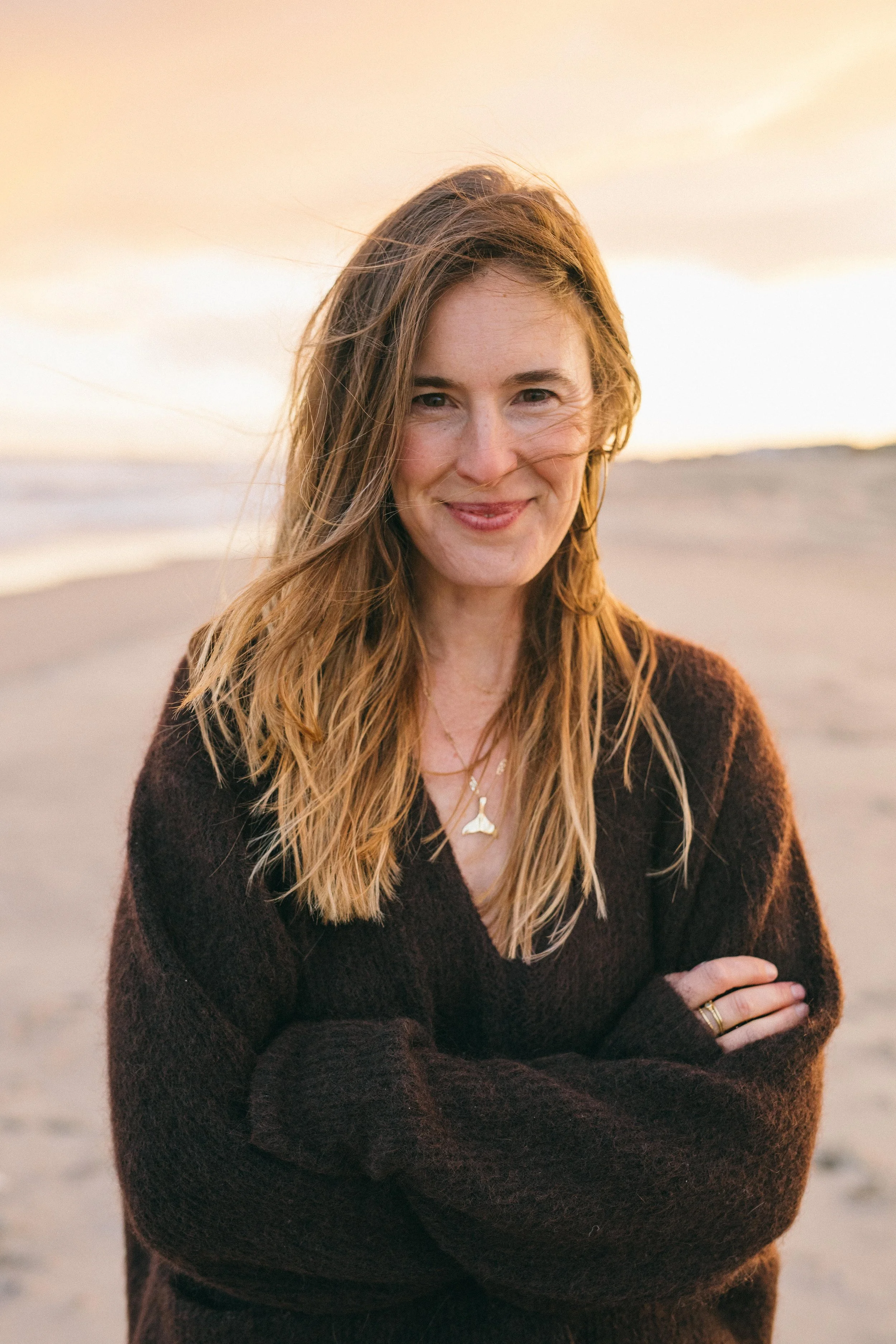 A woman with long, wavy hair smiling with arms crossed, standing on a beach during sunset.