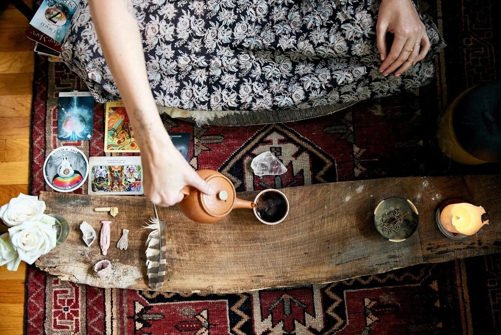 A person sitting on a patterned rug, pouring tea from a teapot into a cup, surrounded by tarot cards, a feather, flowers, candles, and crystals on a wooden table.