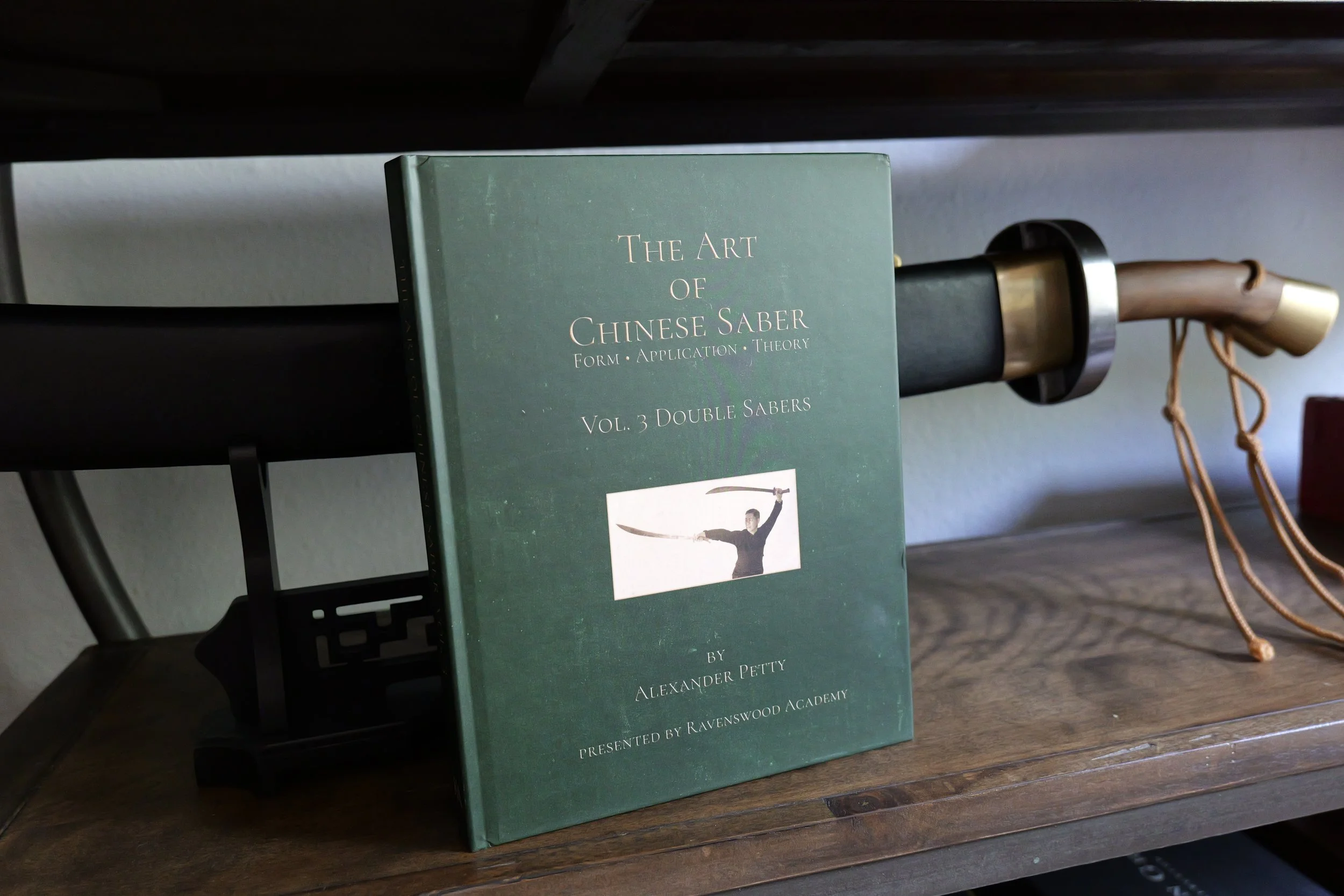 Double Broadswords book on a shelf in front of two Chinese sabers in a sheath.